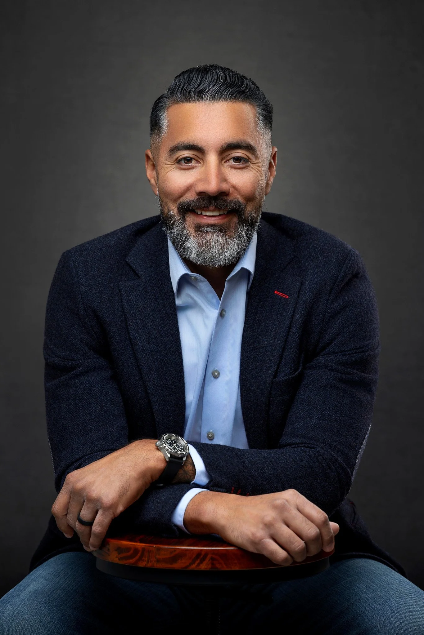 A smiling middle-aged man with a beard, wearing a blue dress shirt and navy blazer, seated against a dark background.