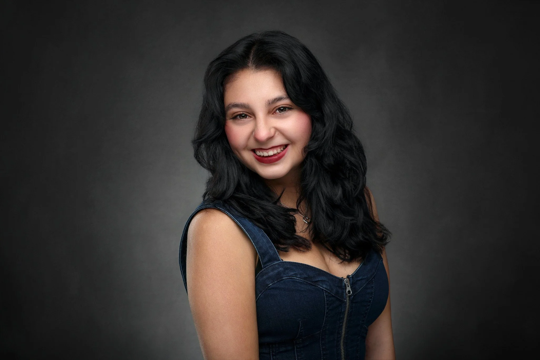 A young woman with long black wavy hair, wearing a sleeveless denim top, smiling against a dark gray background.