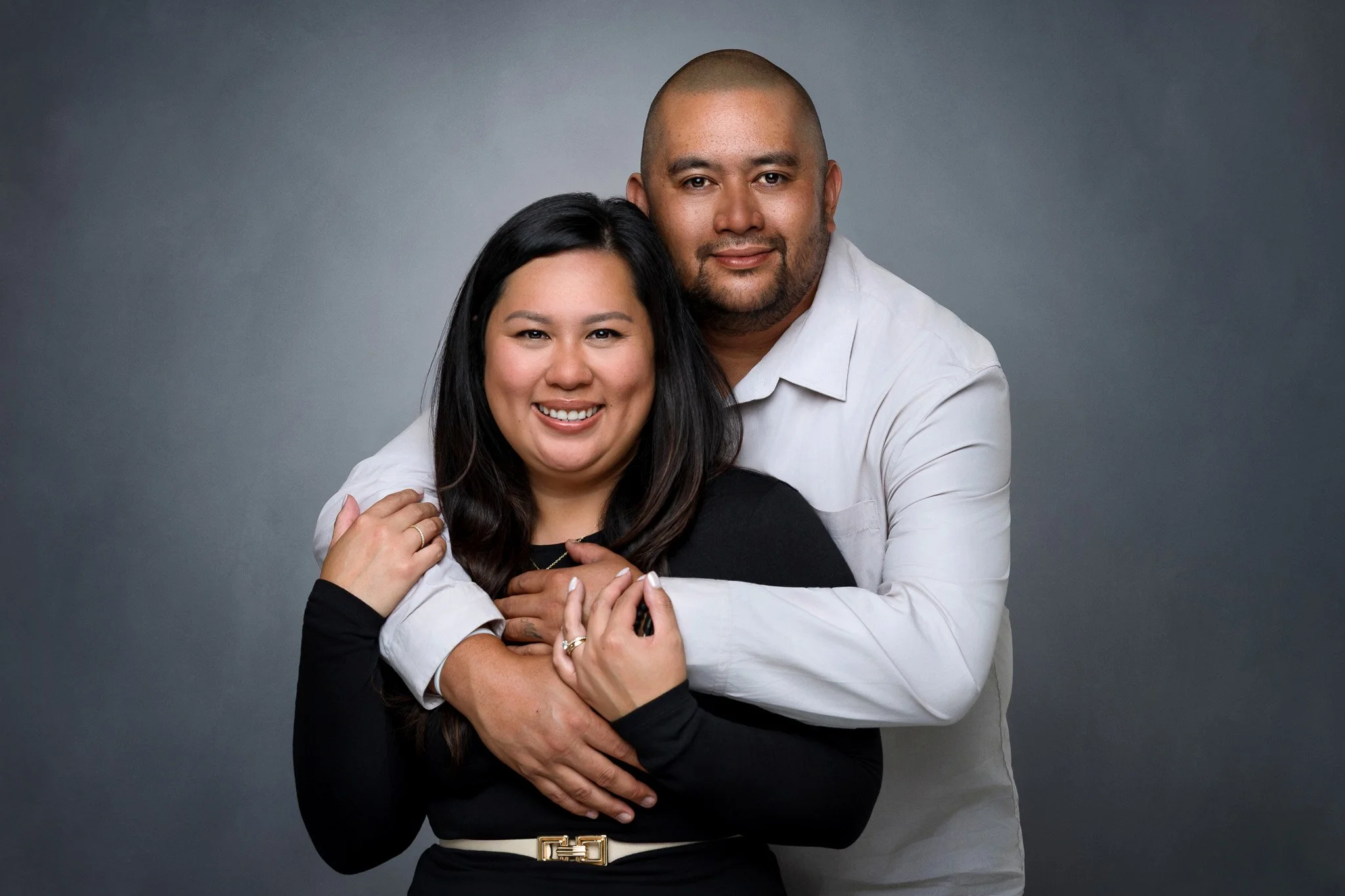 A smiling woman with long dark hair and leather belt, and a man with a shaved head and beard, embracing each other against a gray background.