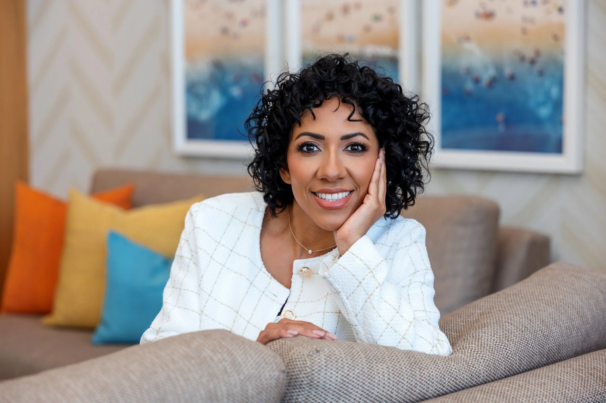 A woman with curly black hair smiling and resting her face on her hand sitting on a beige couch with colorful pillows in a living room.