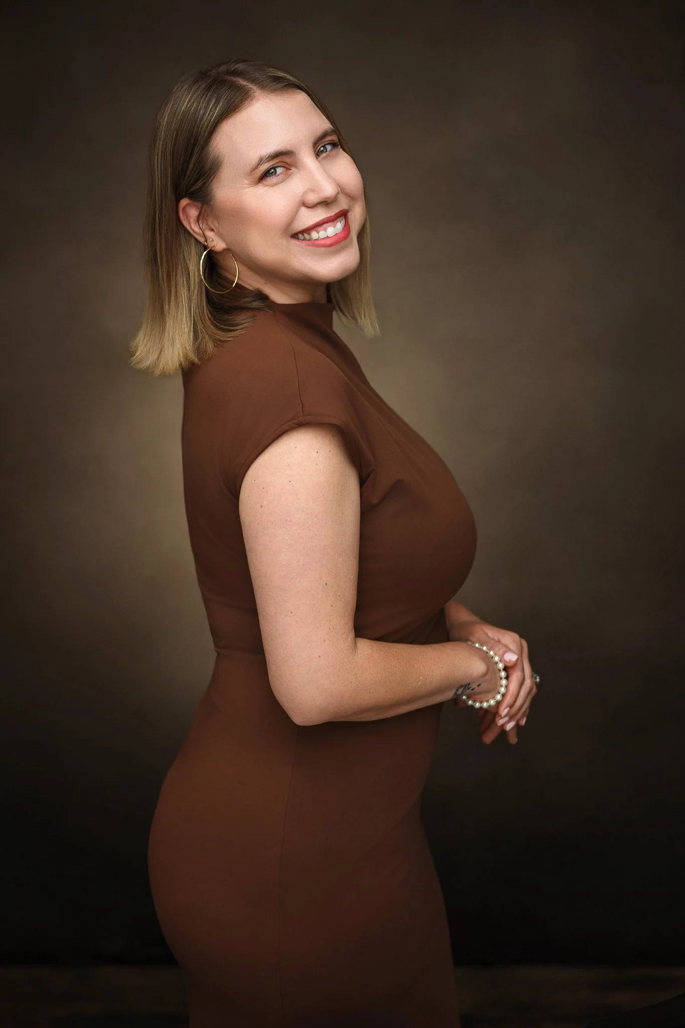 Smiling woman in a brown dress with hoop earrings and bracelet, posing against a neutral background.
