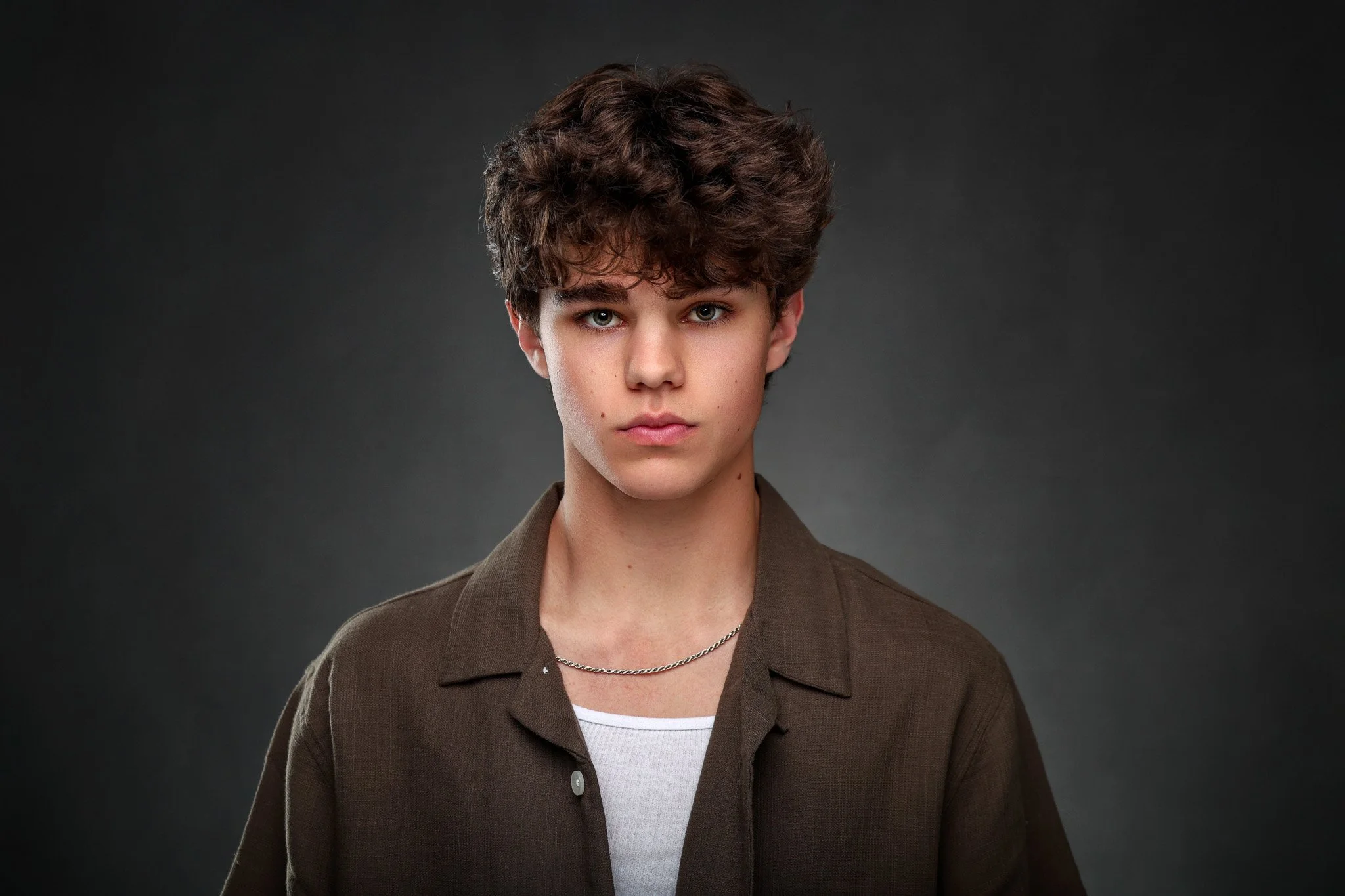 A young person with short, dark, curly hair and blue eyes wearing a brown jacket over a white shirt and a silver chain, against a dark gray background.