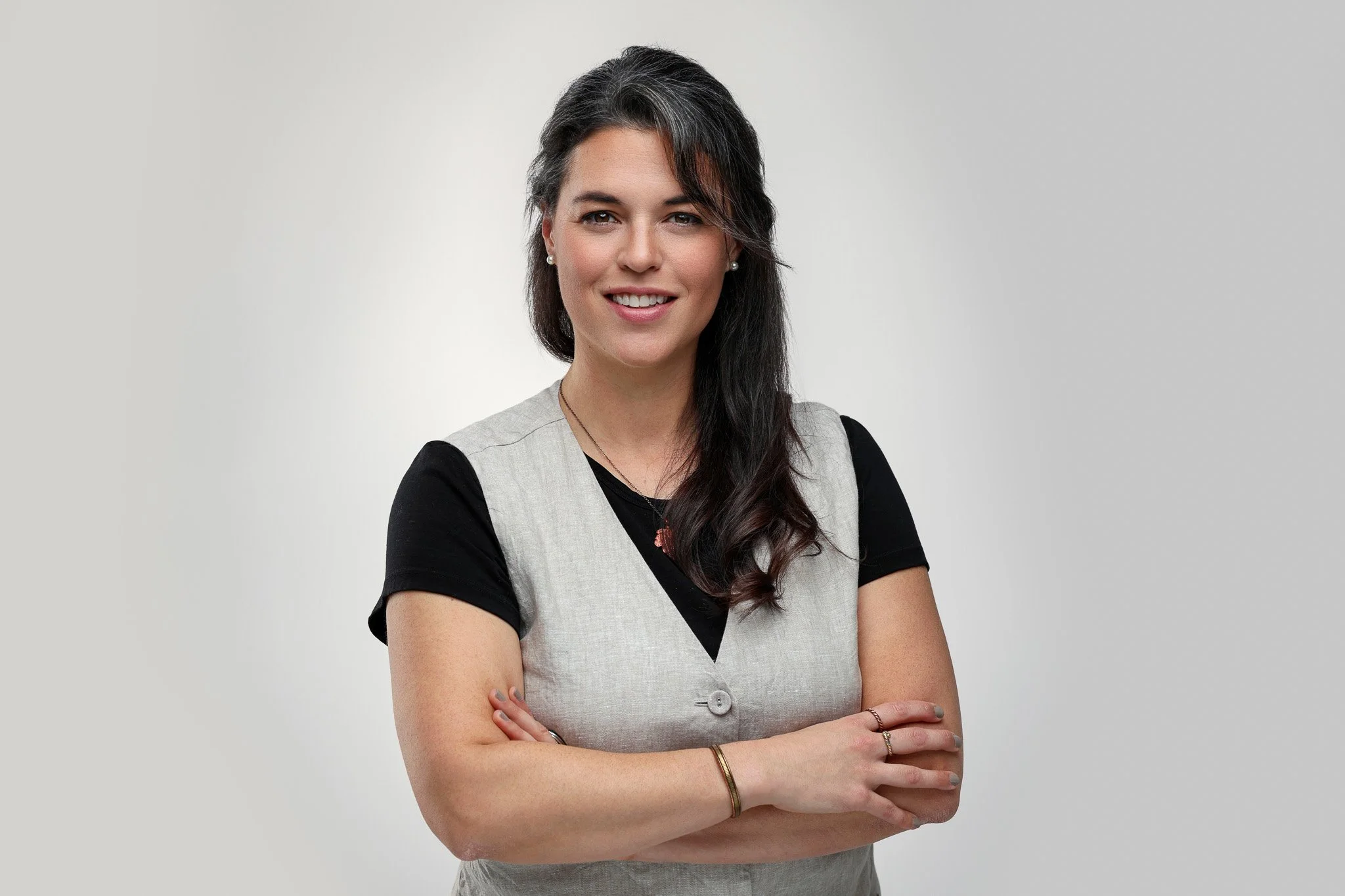 A woman with dark hair, wearing a gray vest over a black shirt, standing with arms crossed against a neutral background.
