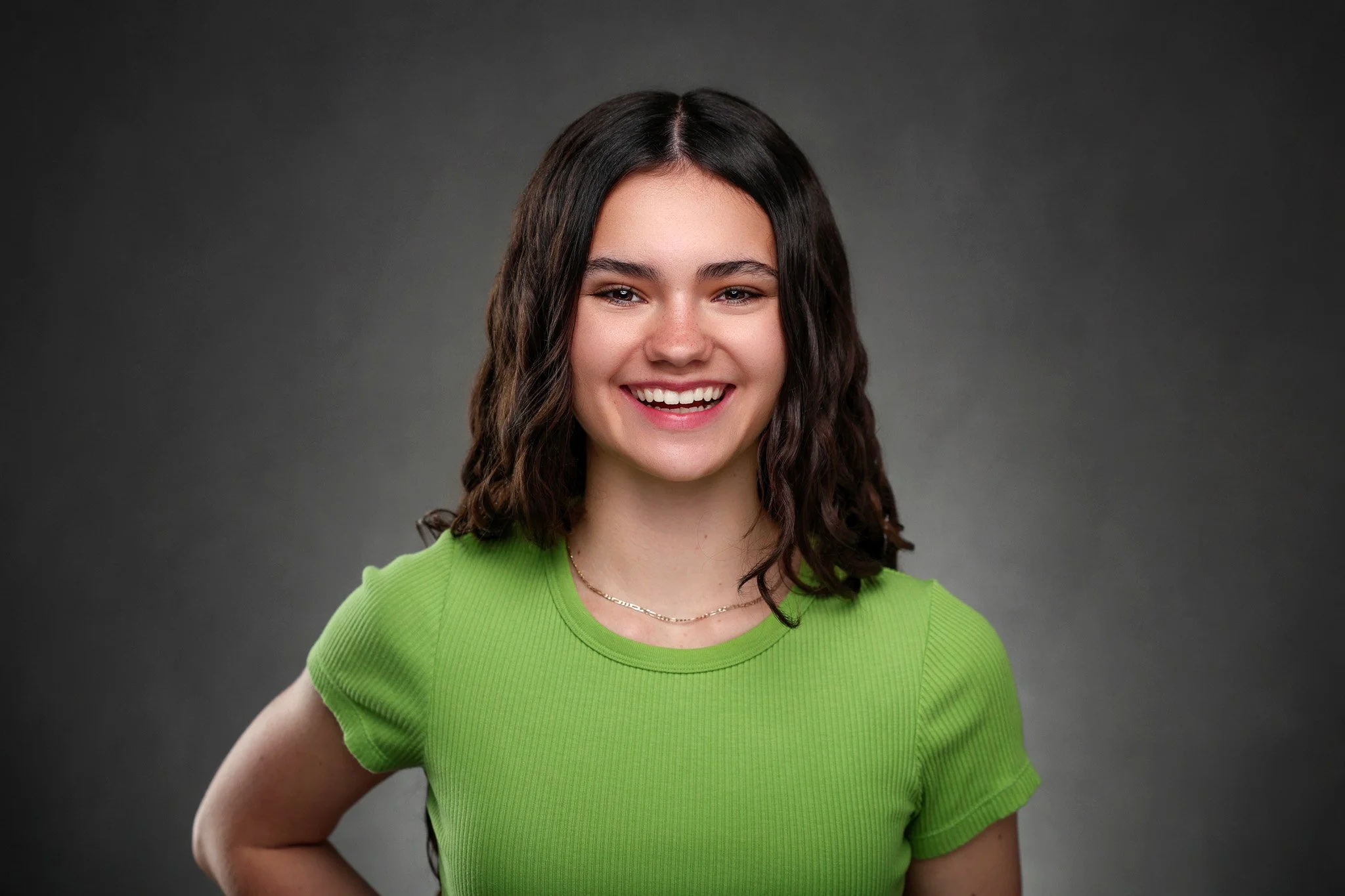 A smiling young woman with long dark brown hair in a lime green shirt against a dark gray background.