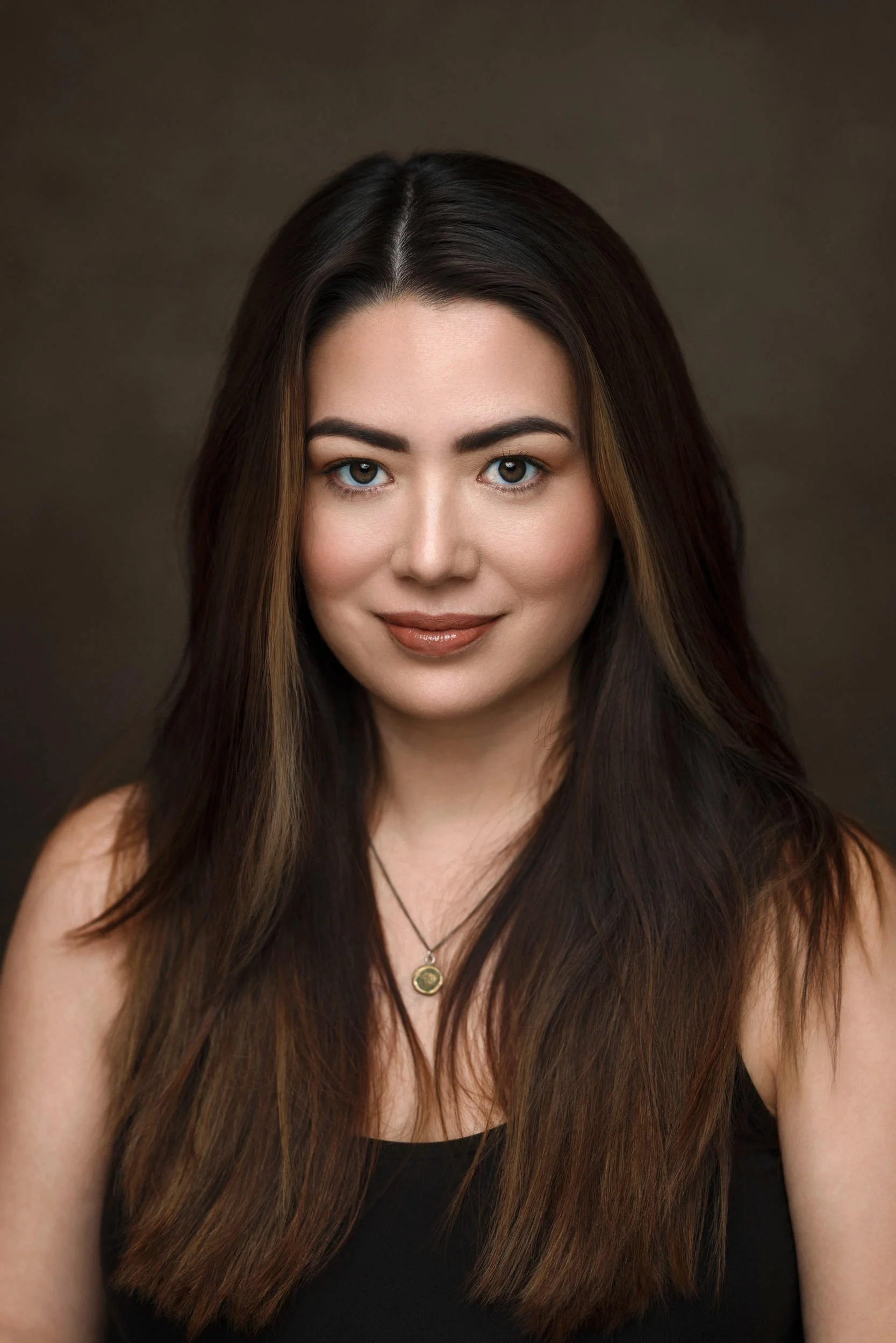 Close-up of a woman with long brown hair, blue eyes, and light makeup, wearing a black top and a necklace with a round pendant, against a dark background.