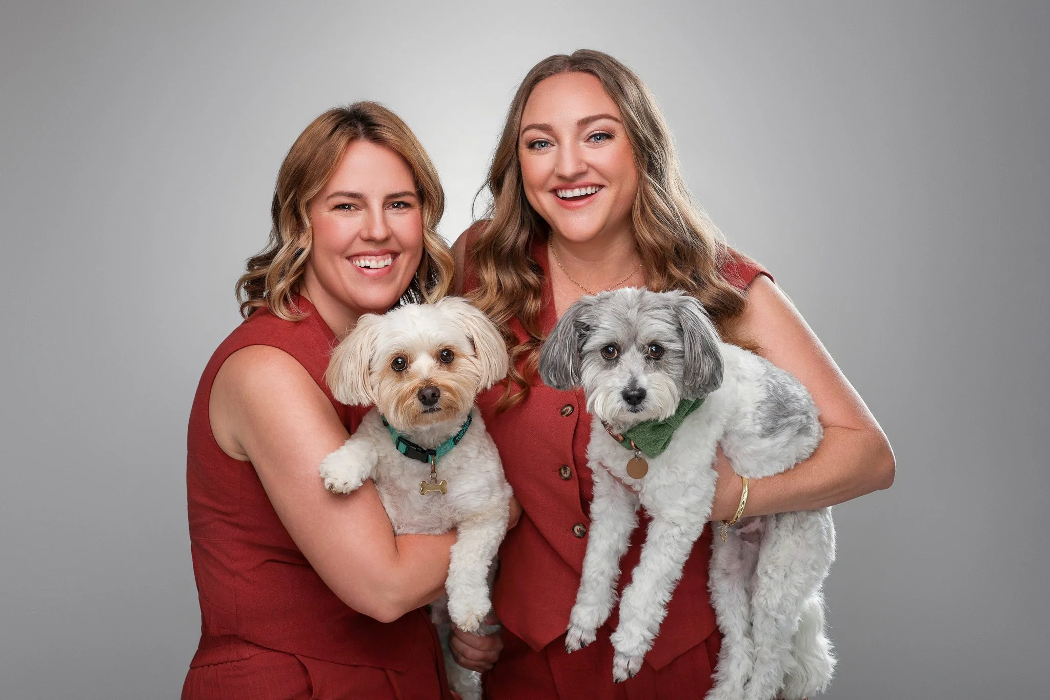 Two smiling women holding their dogs against a plain gray background.