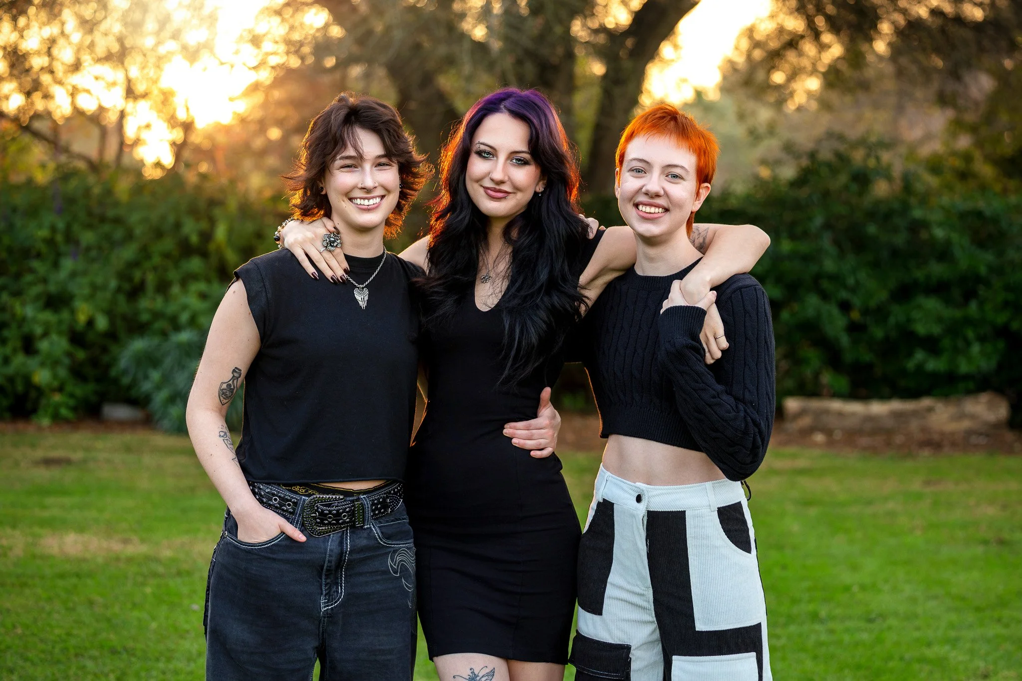 Three young women standing close together outdoors during sunset, smiling at the camera with arms around each other, in front of trees and greenery.