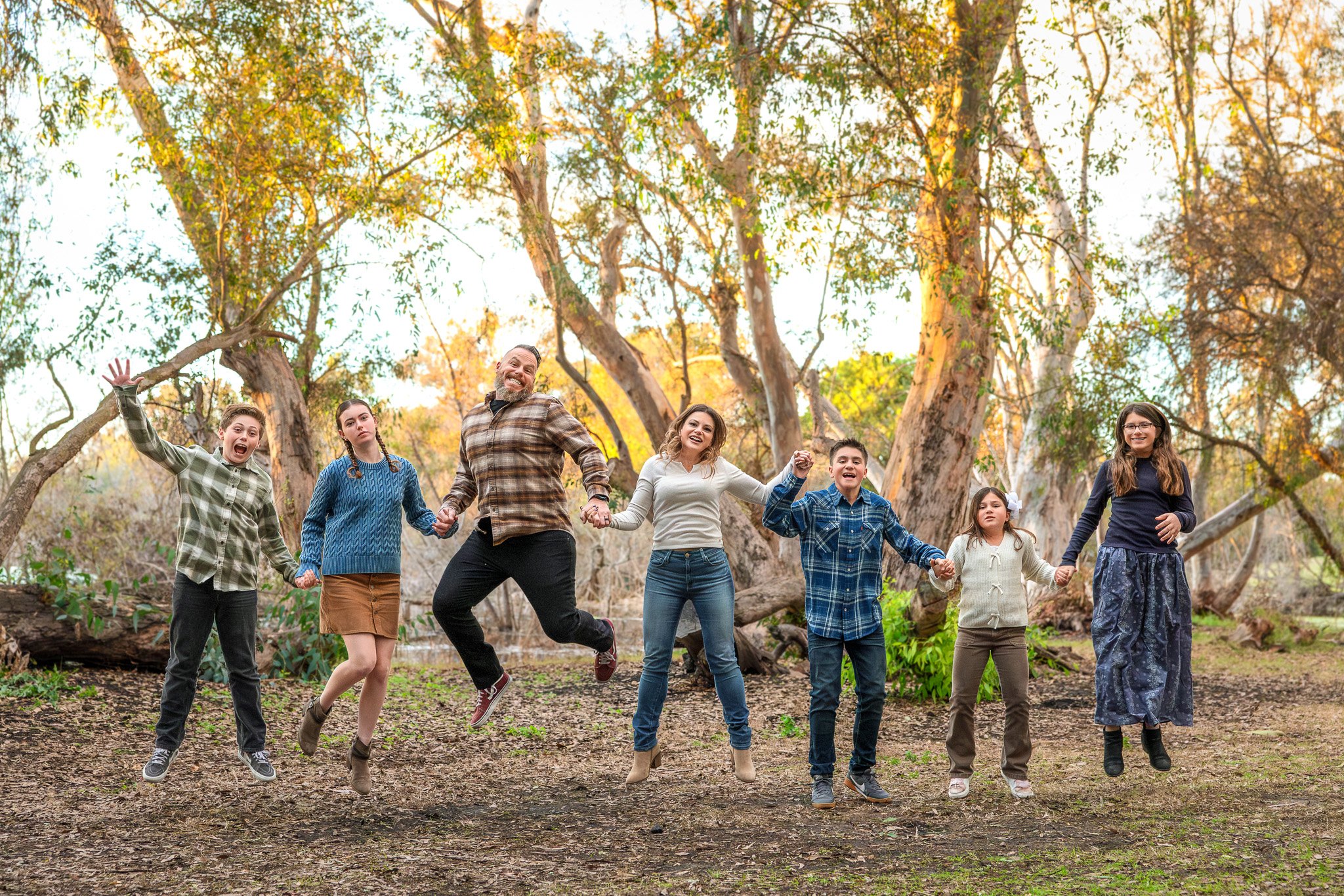 A family of eight kids and two adults jumping hand-in-hand outdoors in a wooded area during autumn.