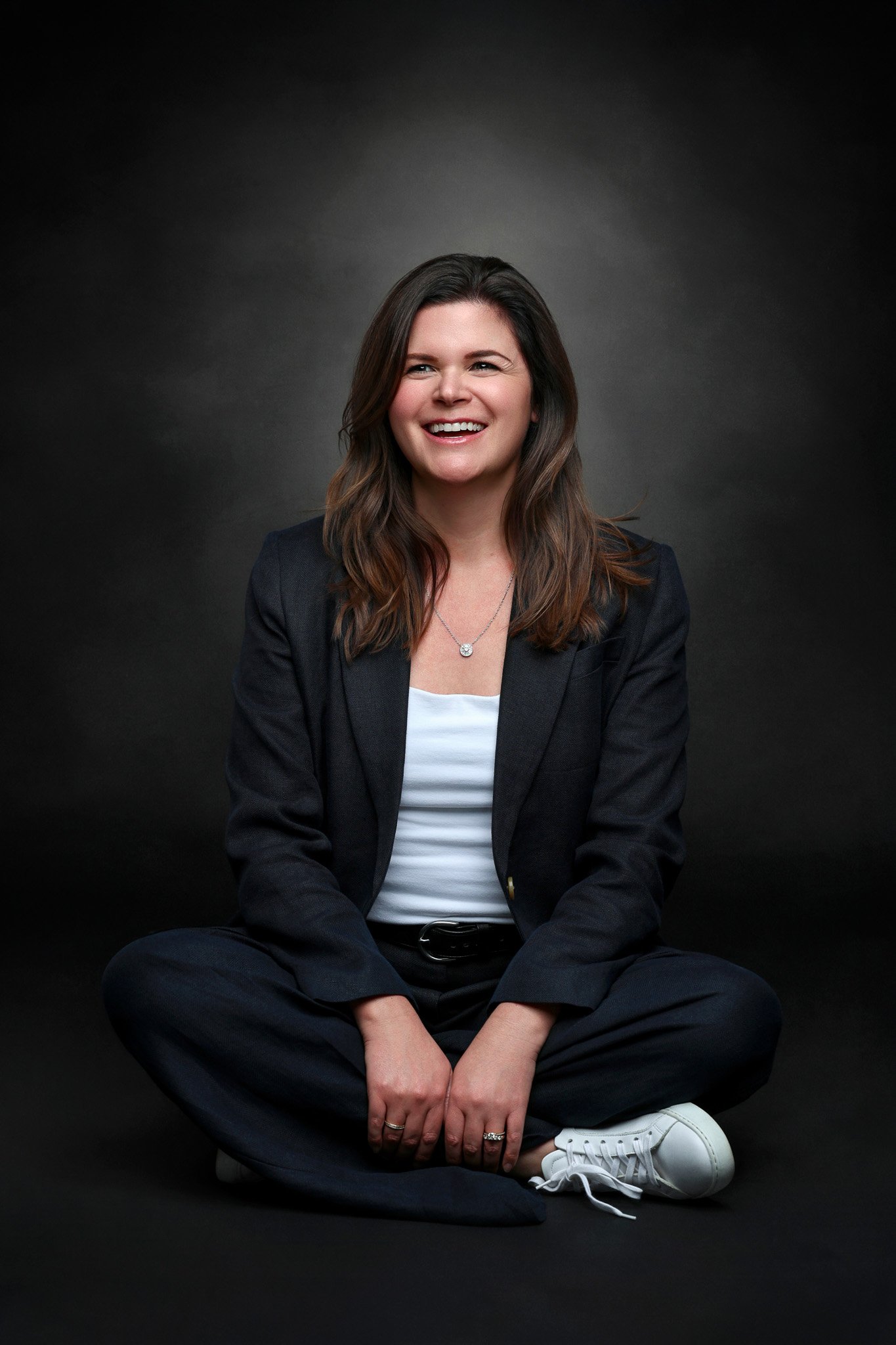 A woman with brown hair, wearing a black blazer, white shirt, and sneakers, sitting cross-legged on a dark floor against a dark background, smiling.