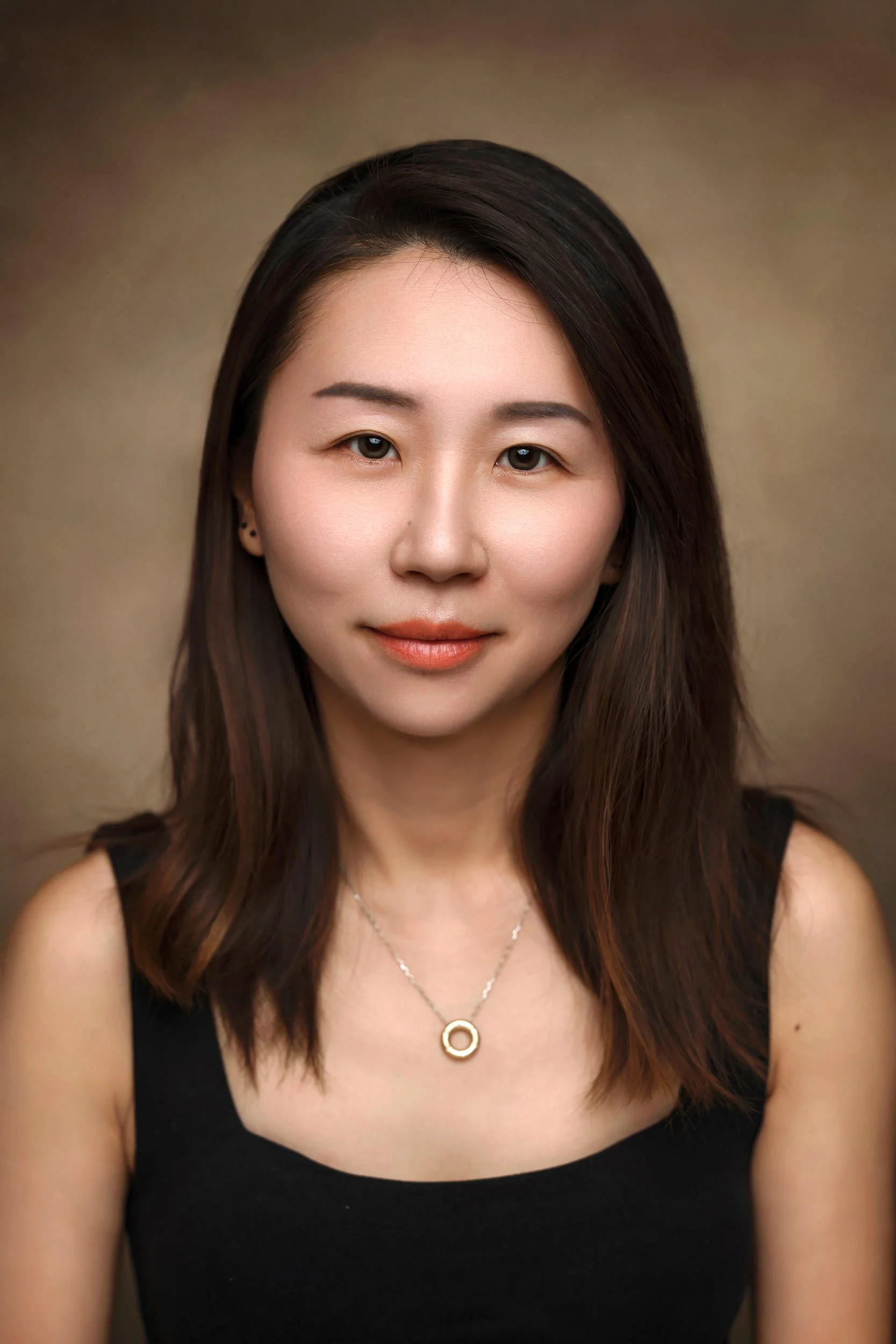 Close-up portrait of a woman with dark brown, shoulder-length hair, wearing a black sleeveless top, a necklace with a circular pendant, and small black earrings, against a neutral background.