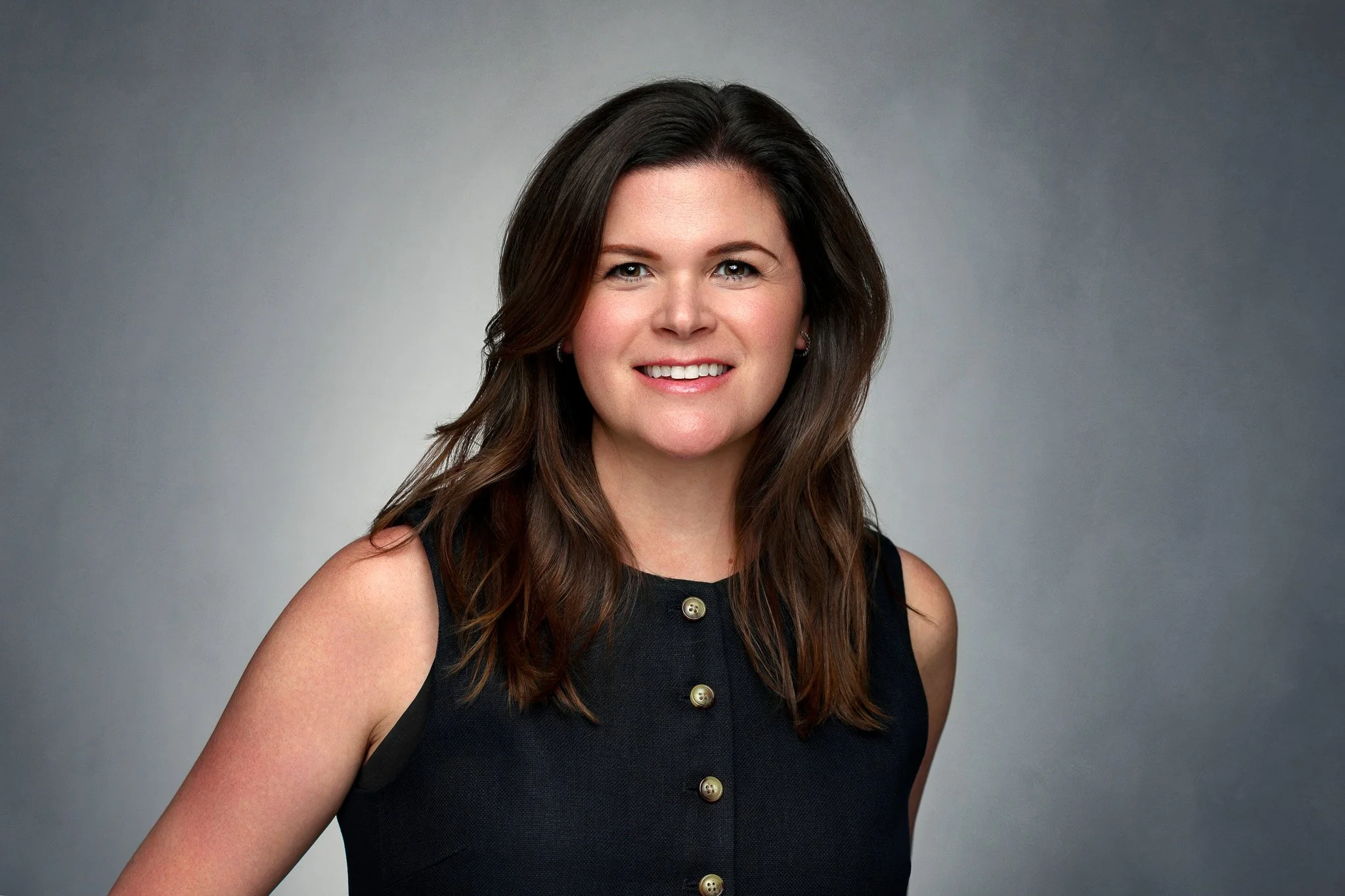 A young woman with brown hair smiling, wearing a sleeveless black top with gold buttons, against a gray background.