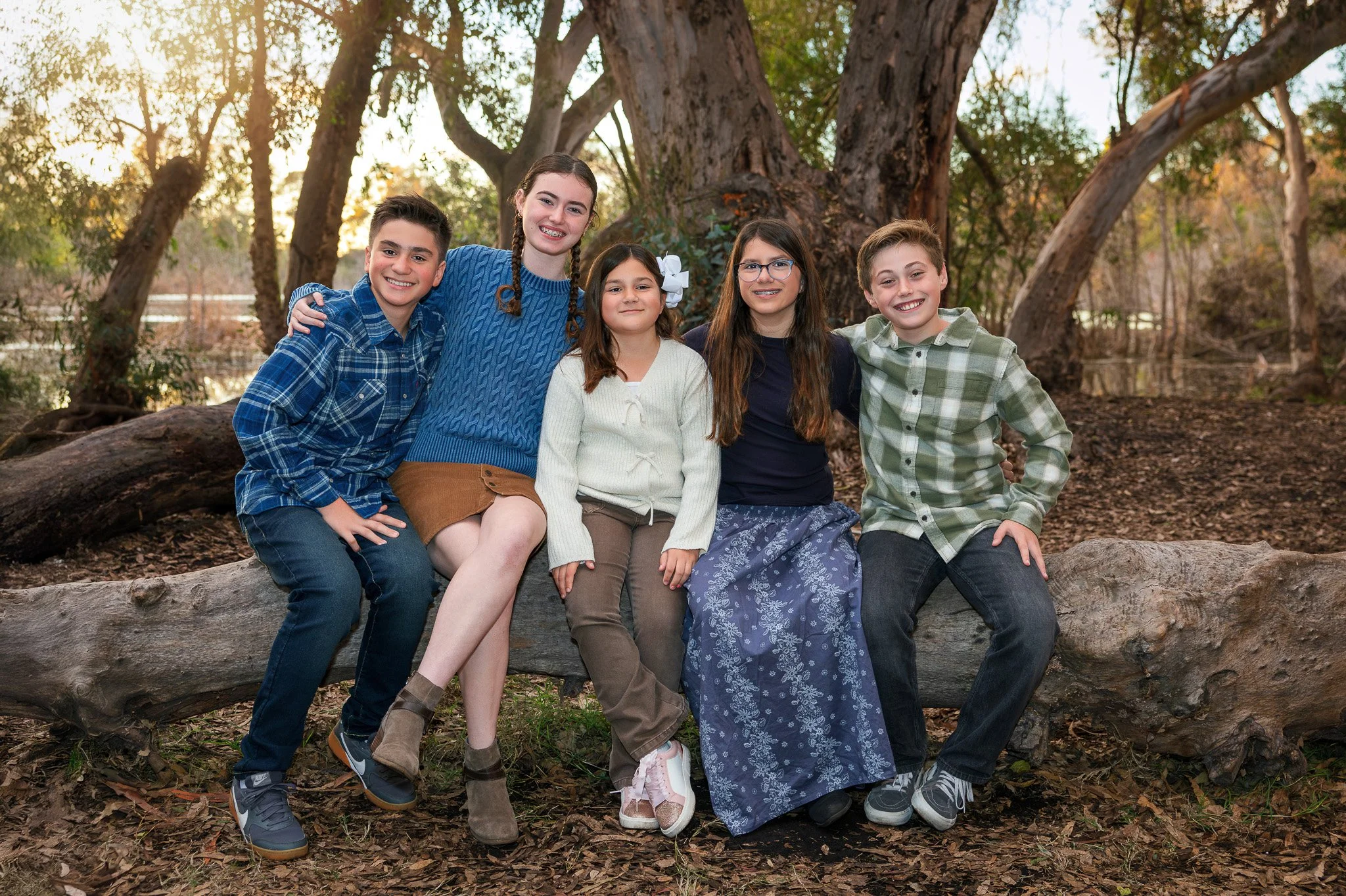 Group of five children sitting on a fallen log outdoors, with trees and a body of water in the background, during sunset.