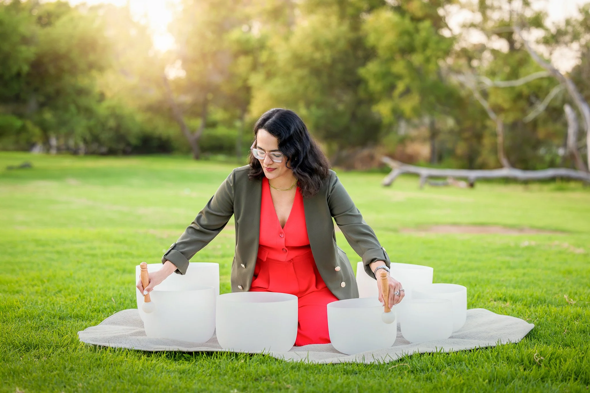Woman in red dress and green blazer playing singing bowls outdoors on a grassy field with trees and sunlight in the background.