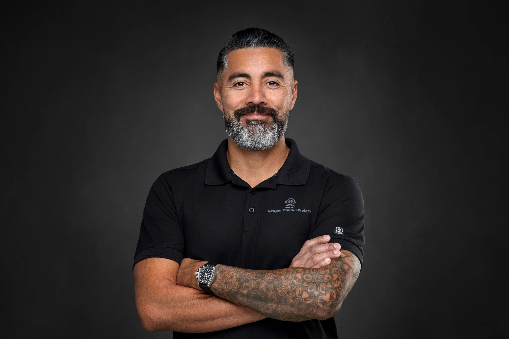 A man with a beard and mustache, wearing a black polo shirt with a logo and text, standing with arms crossed against a dark background.