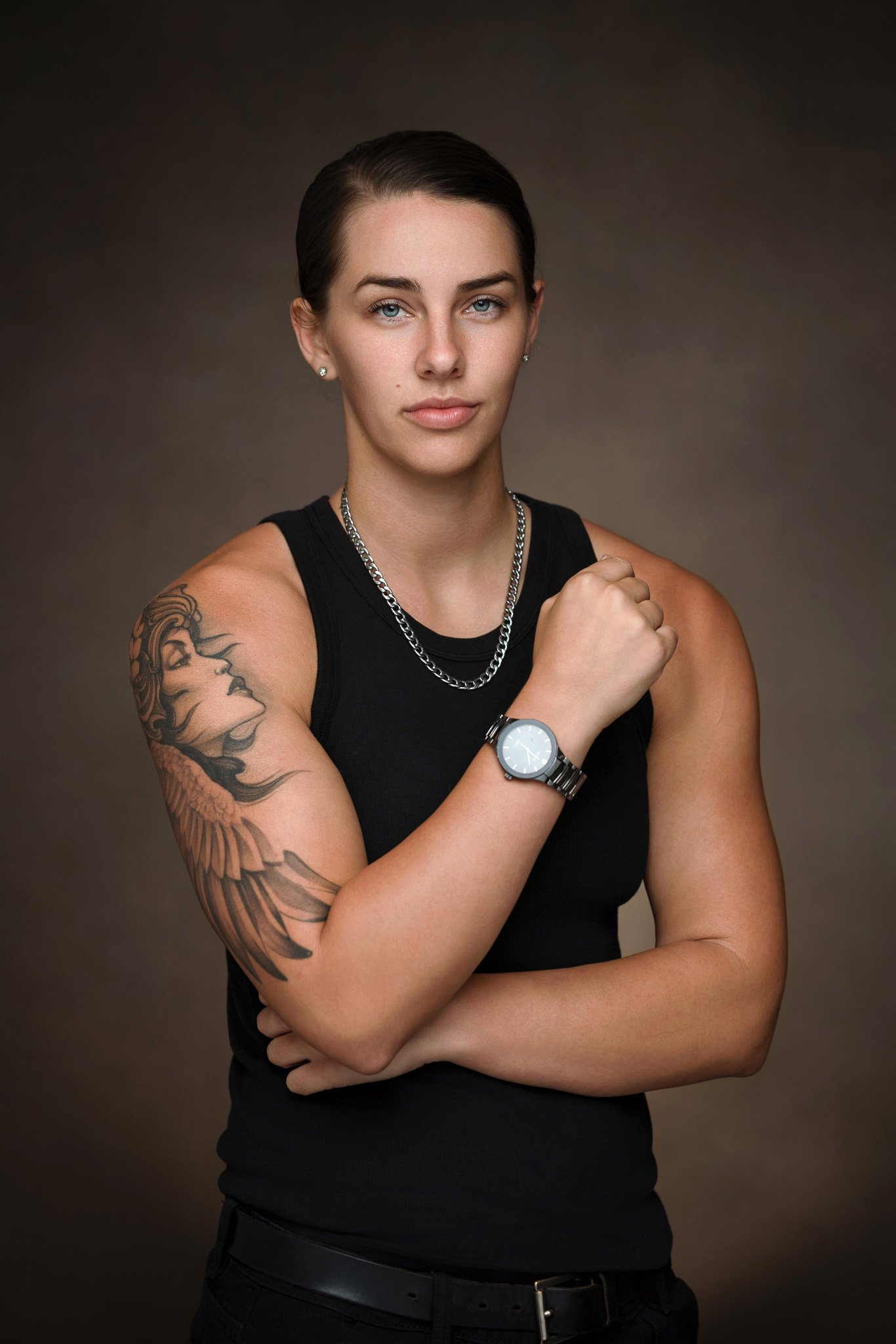 A woman with short dark hair, blue eyes, and tattoos on her arm, wearing a black sleeveless top, silver chain necklace, and watch, standing against a dark gradient background.