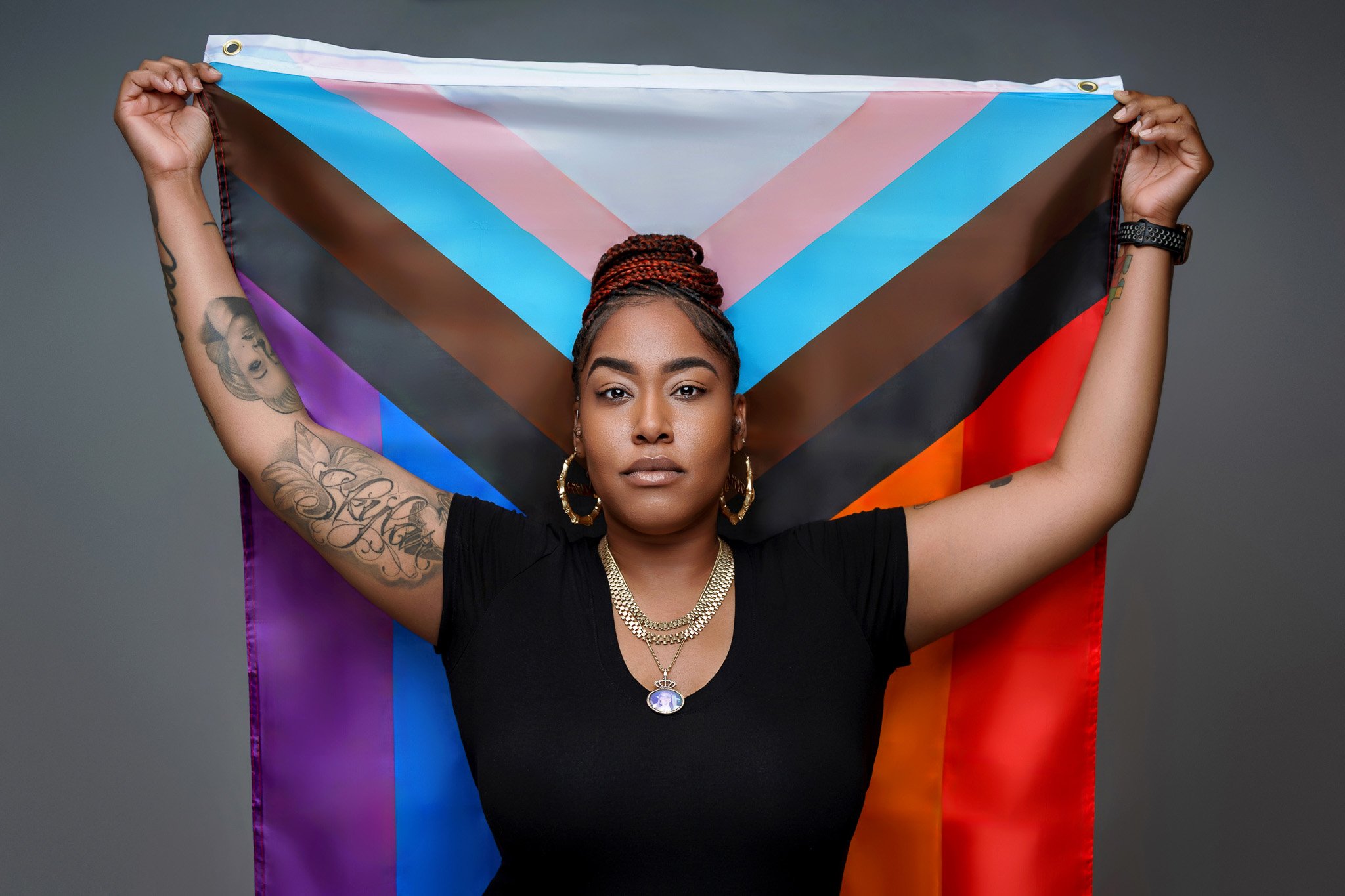 A woman with tattoos on her arms is holding up a transgender pride flag behind her shoulders. She has braided hair, gold hoop earrings, a layered gold necklace, and is wearing a black shirt against a gray background.