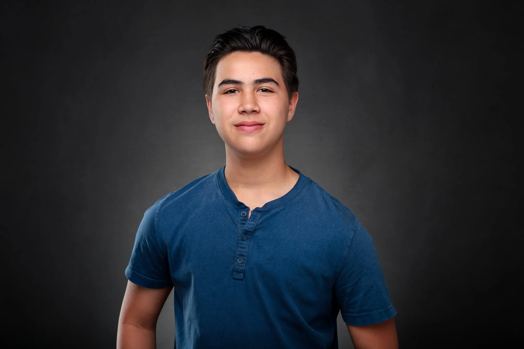 A young man with short dark hair and fair skin wears a blue shirt against a dark gray background.