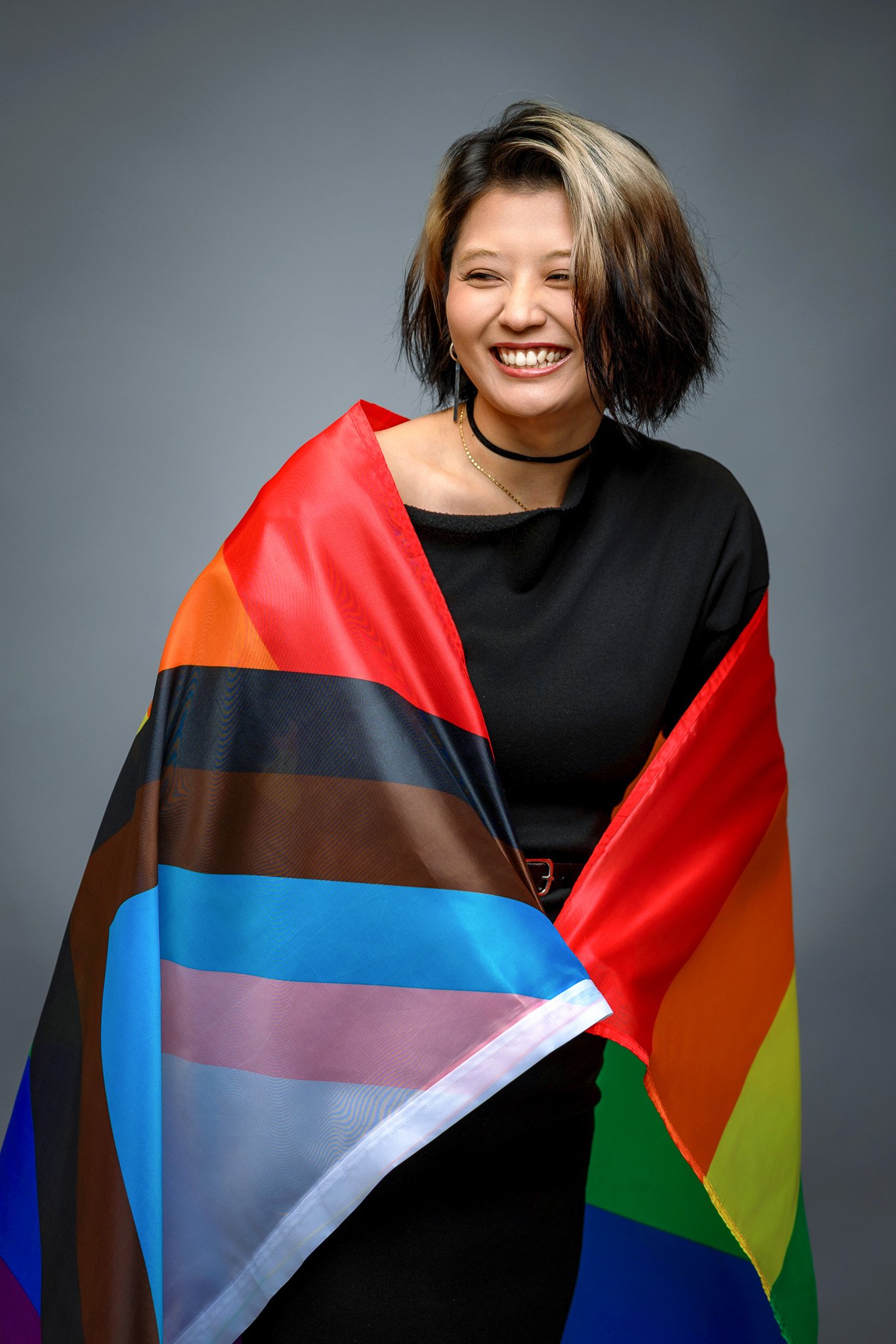 A woman with short, wavy hair laughing, draped in a rainbow pride flag, standing against a gray background.
