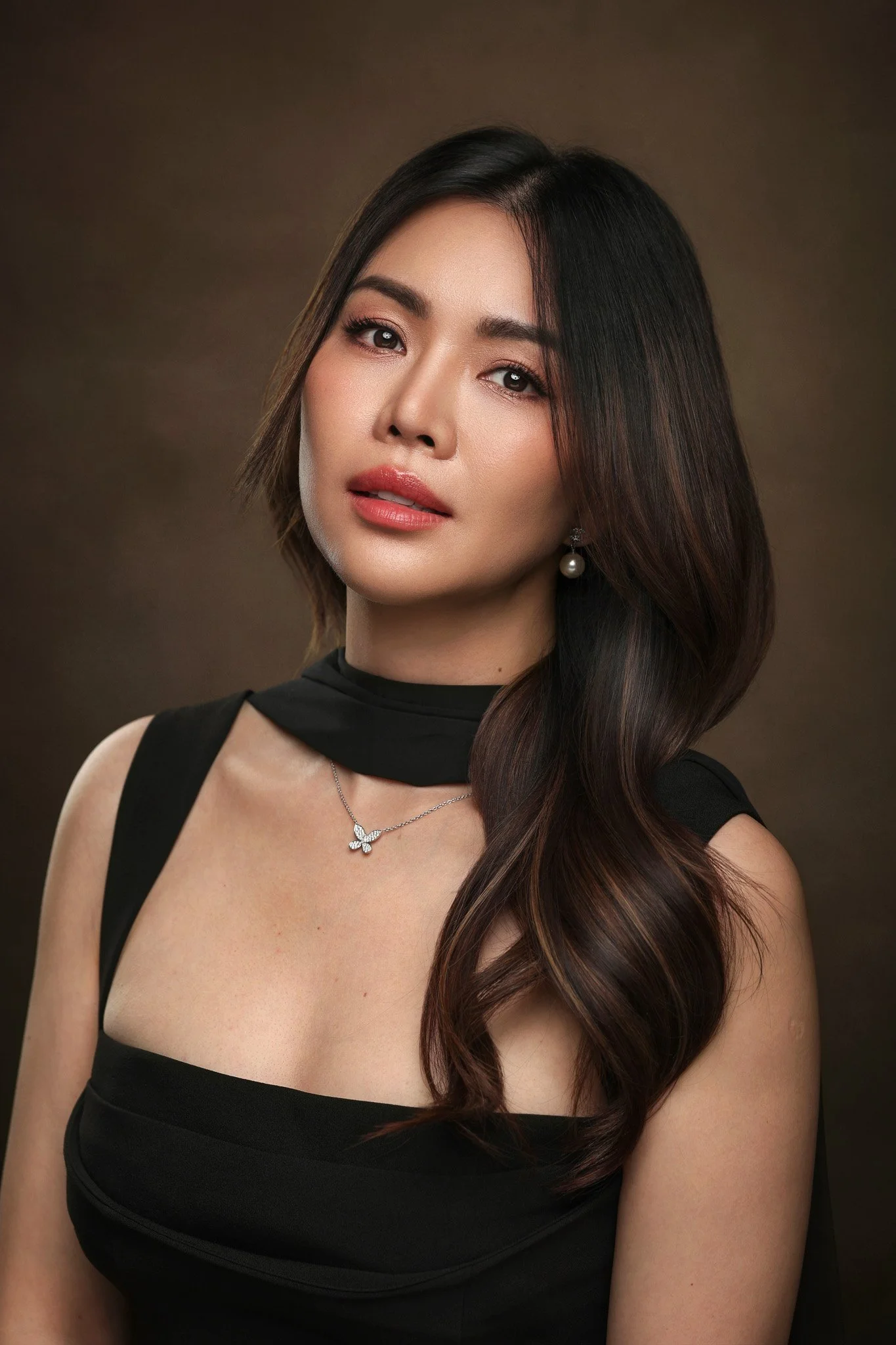 Portrait of a woman with long dark hair, wearing a black dress and pearl earrings, against a dark background.