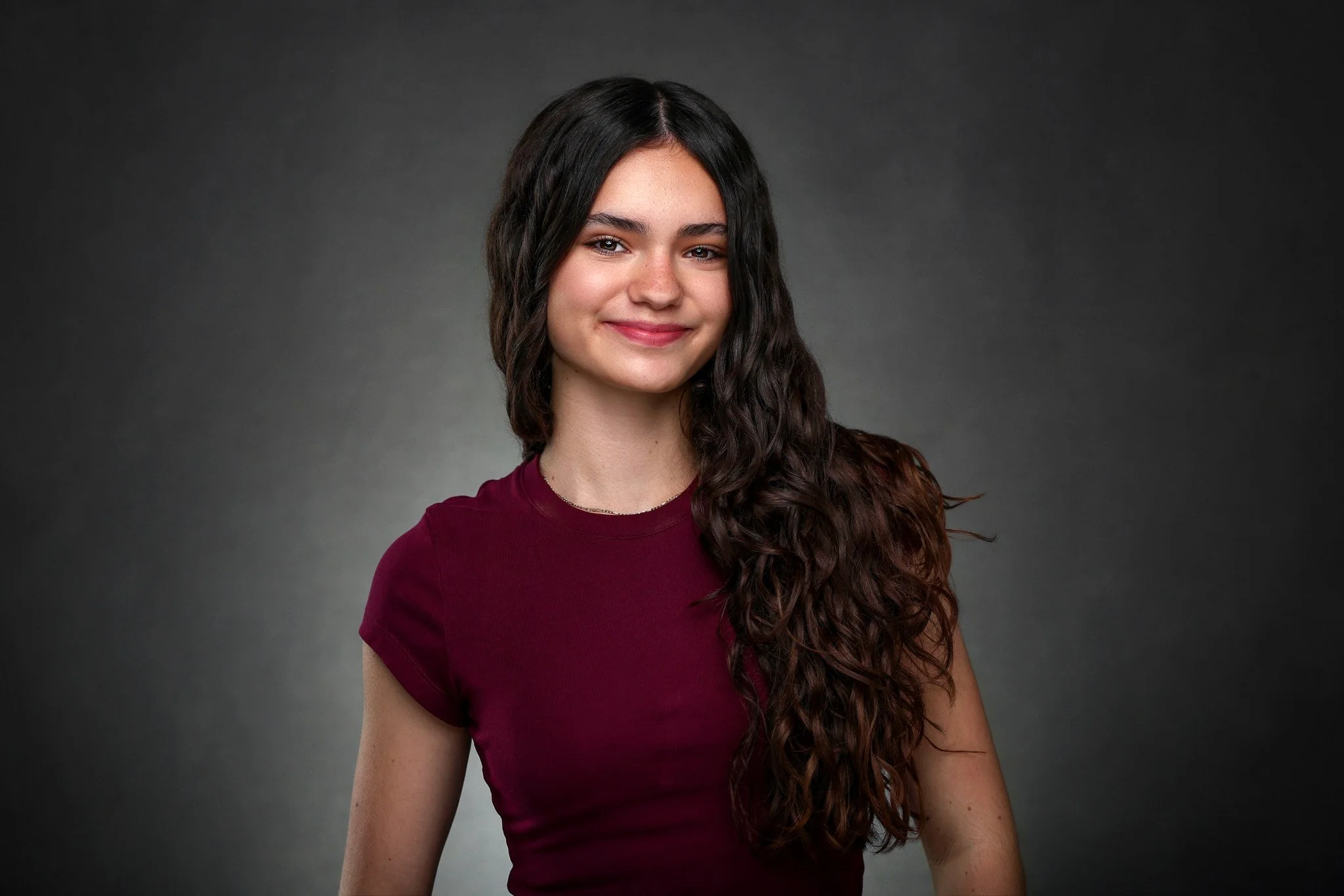 A young woman with long, dark, wavy hair wearing a burgundy top standing against a dark gray background.