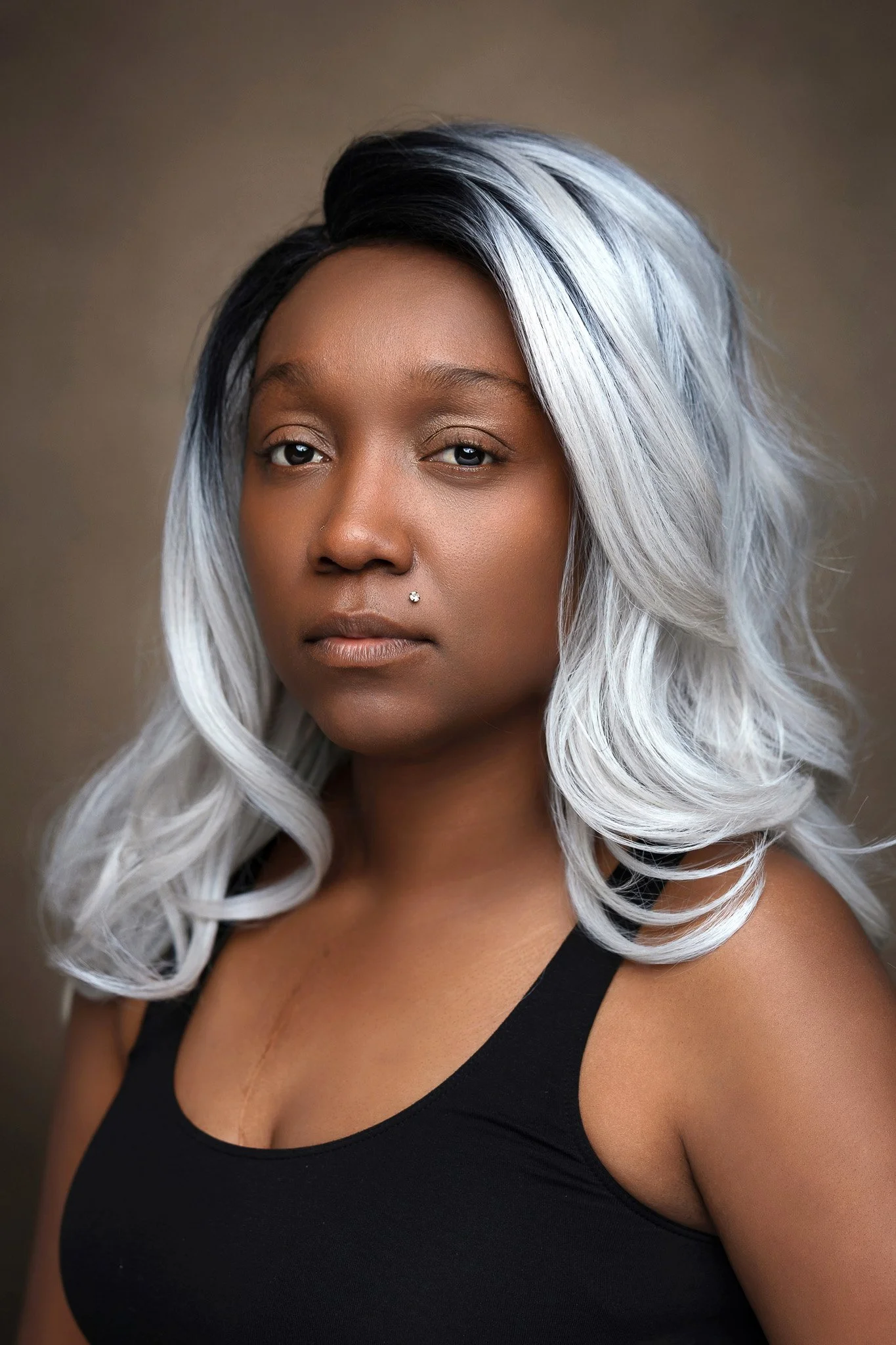Close-up of woman with black and silver hair, nose piercing, wearing a black tank top, against a neutral background.
