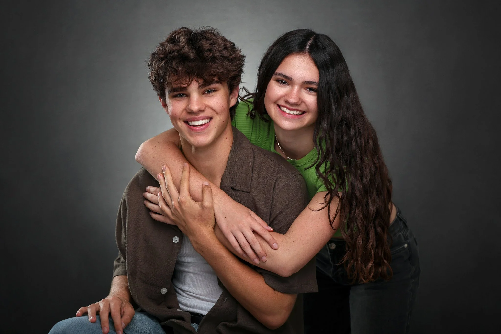 A young man and woman smiling, the woman hugging the man from behind, both looking at the camera, against a neutral gray background.