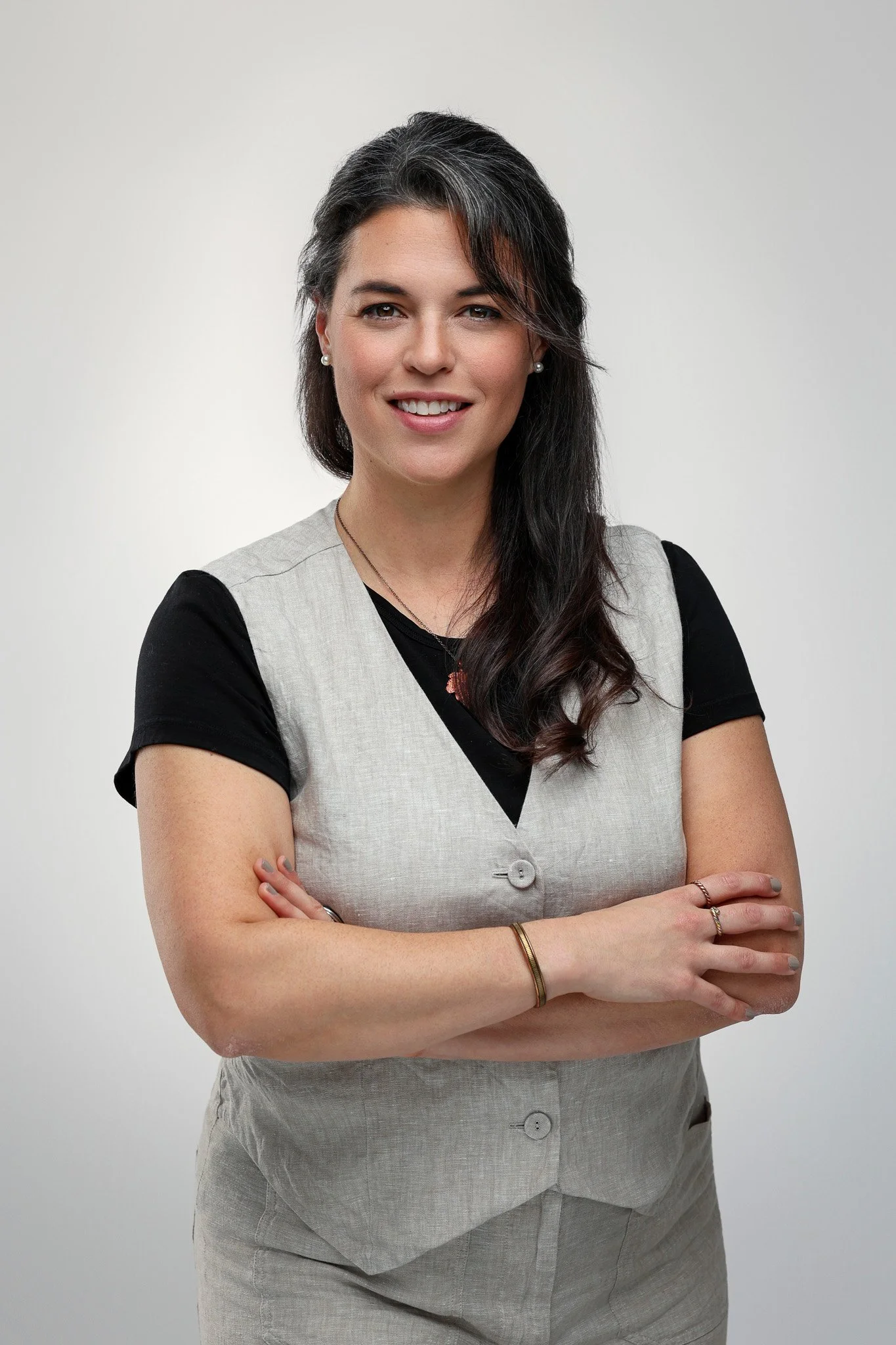 A woman with dark hair, smiling with arms crossed, wearing a black shirt under a gray vest, standing against a plain light background.