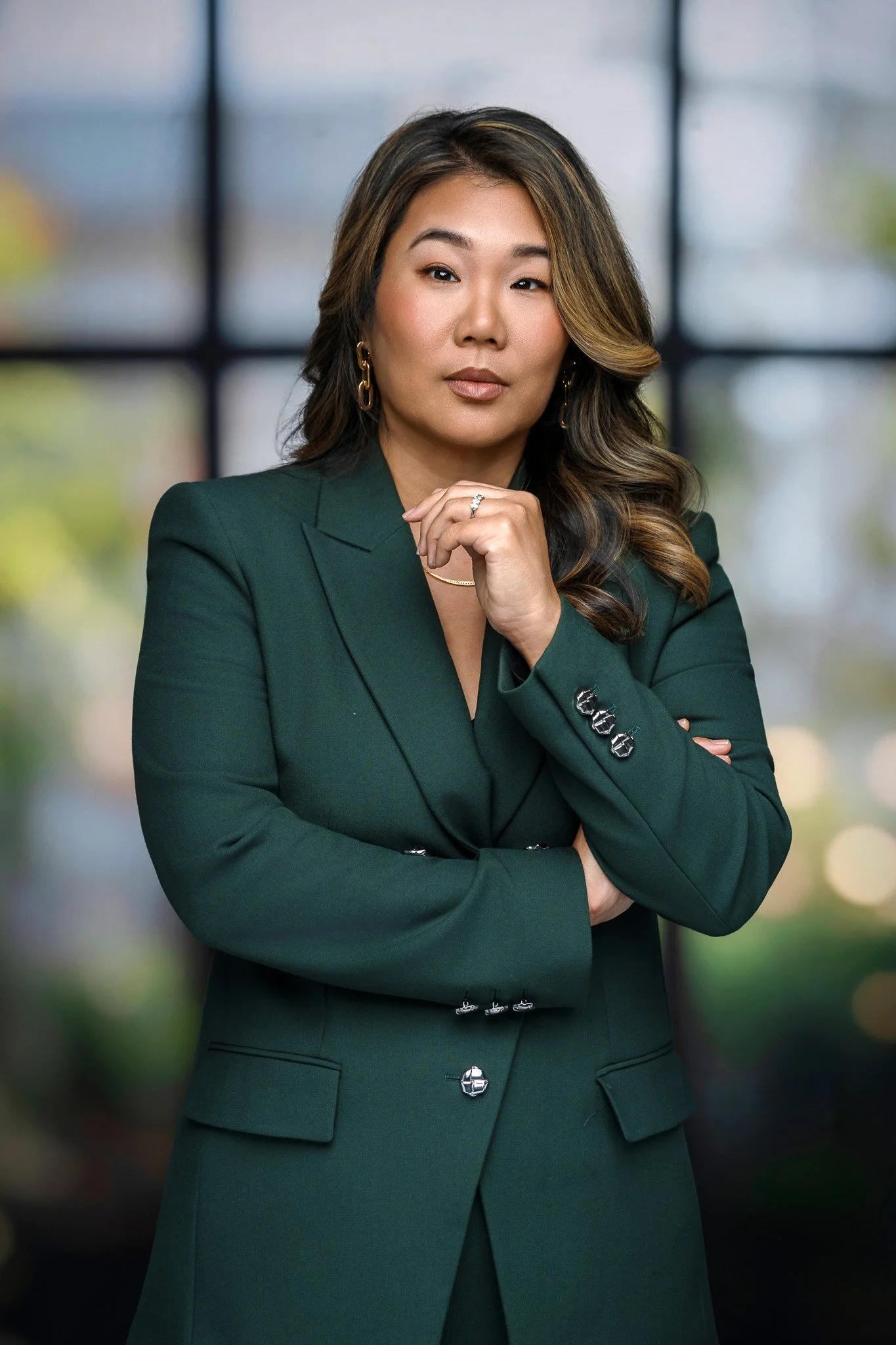 Professional woman in a dark green blazer with wavy hair, standing with crossed arms and a contemplative expression in front of a window with blurred outdoor scenery.