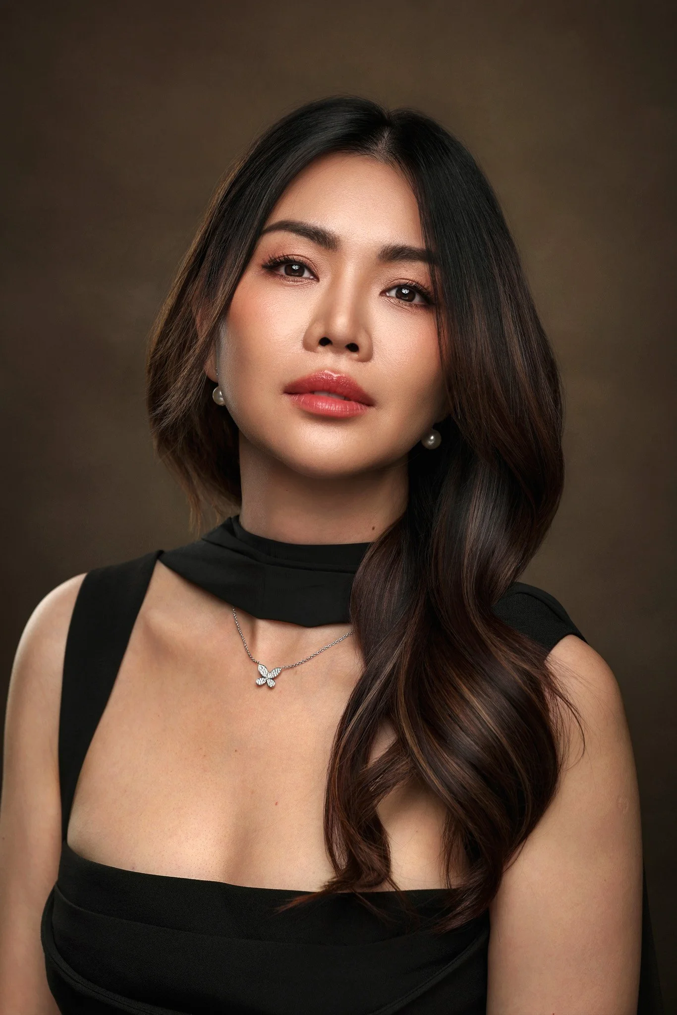 A portrait of a woman with long, wavy dark hair, wearing pearl earrings, a butterfly-shaped pendant necklace, and a black top with cut-out shoulders, against a dark brown background.