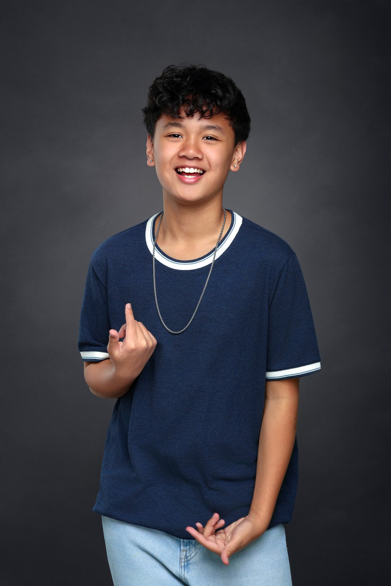 A smiling young person with short curly hair, wearing a dark blue t-shirt with white trim and light-wash jeans, standing against a dark gray background.