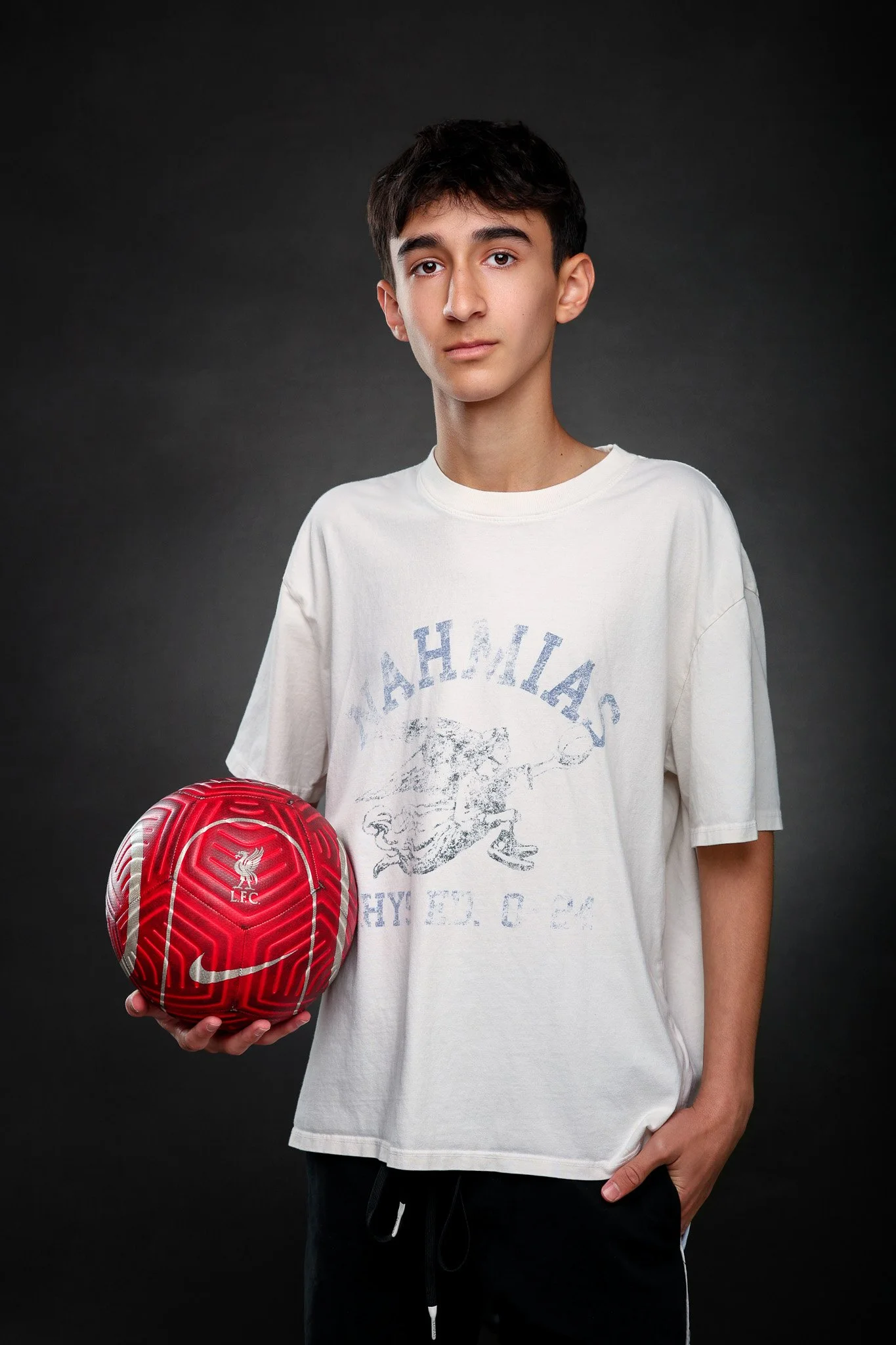 Young male football player holding a red Liverpool Football Club soccer ball, wearing a loose white T-shirt against a dark background.