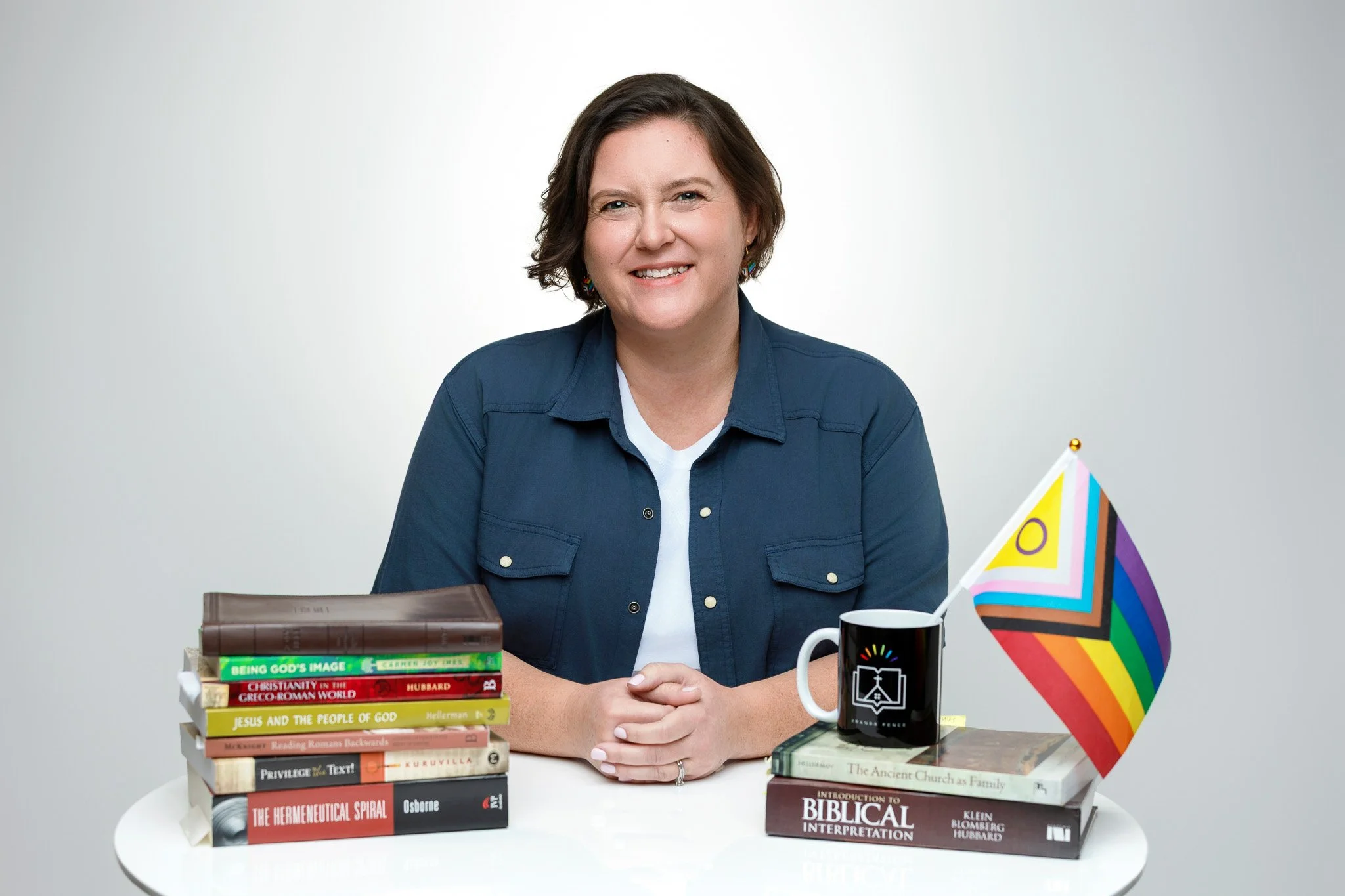 A woman with short brown hair smiling, sitting at a white table with stacks of religious and biblical books, a black mug, and a rainbow pride flag.