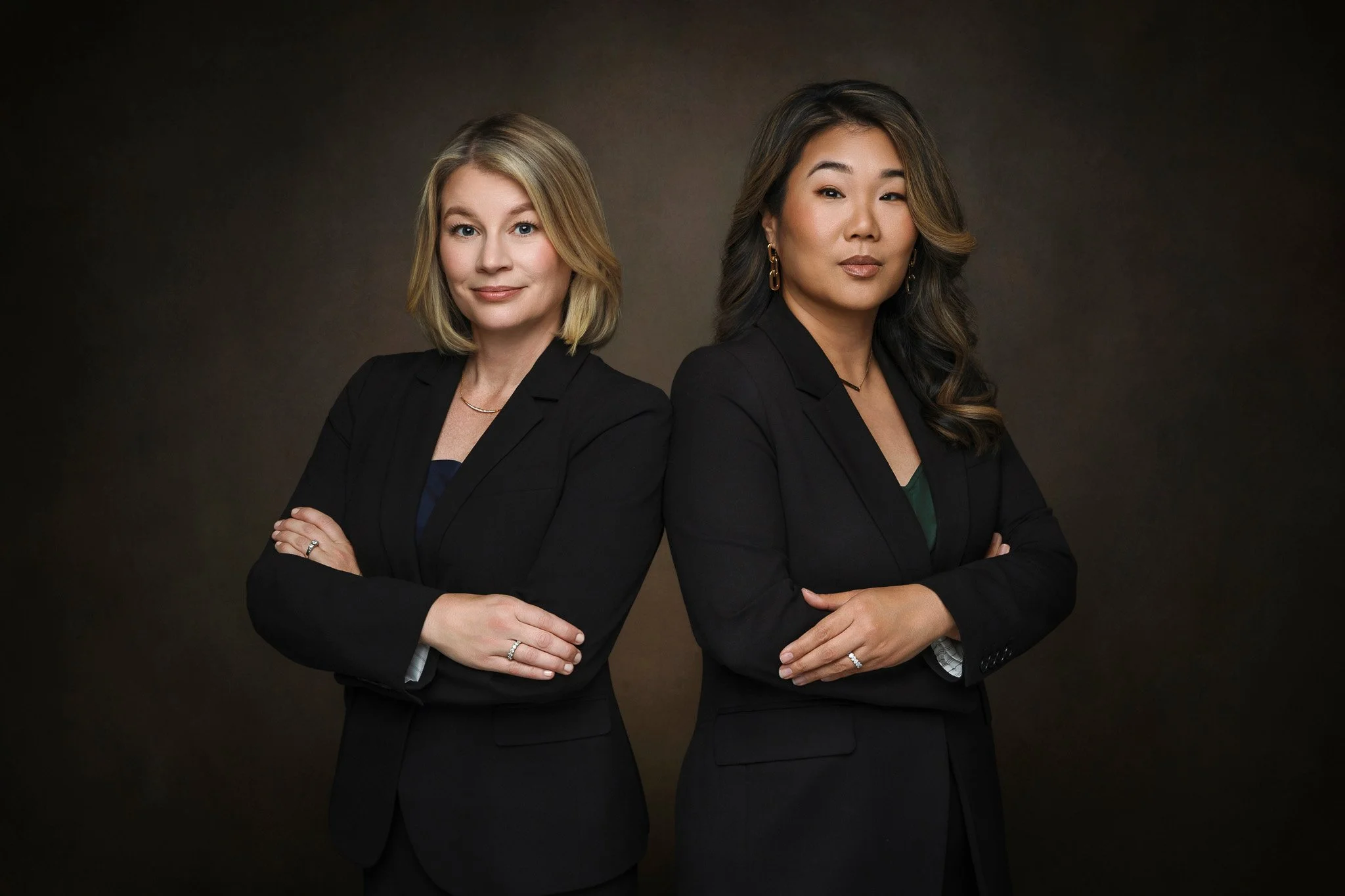 Two women in black business suits standing with their arms crossed against a dark background.