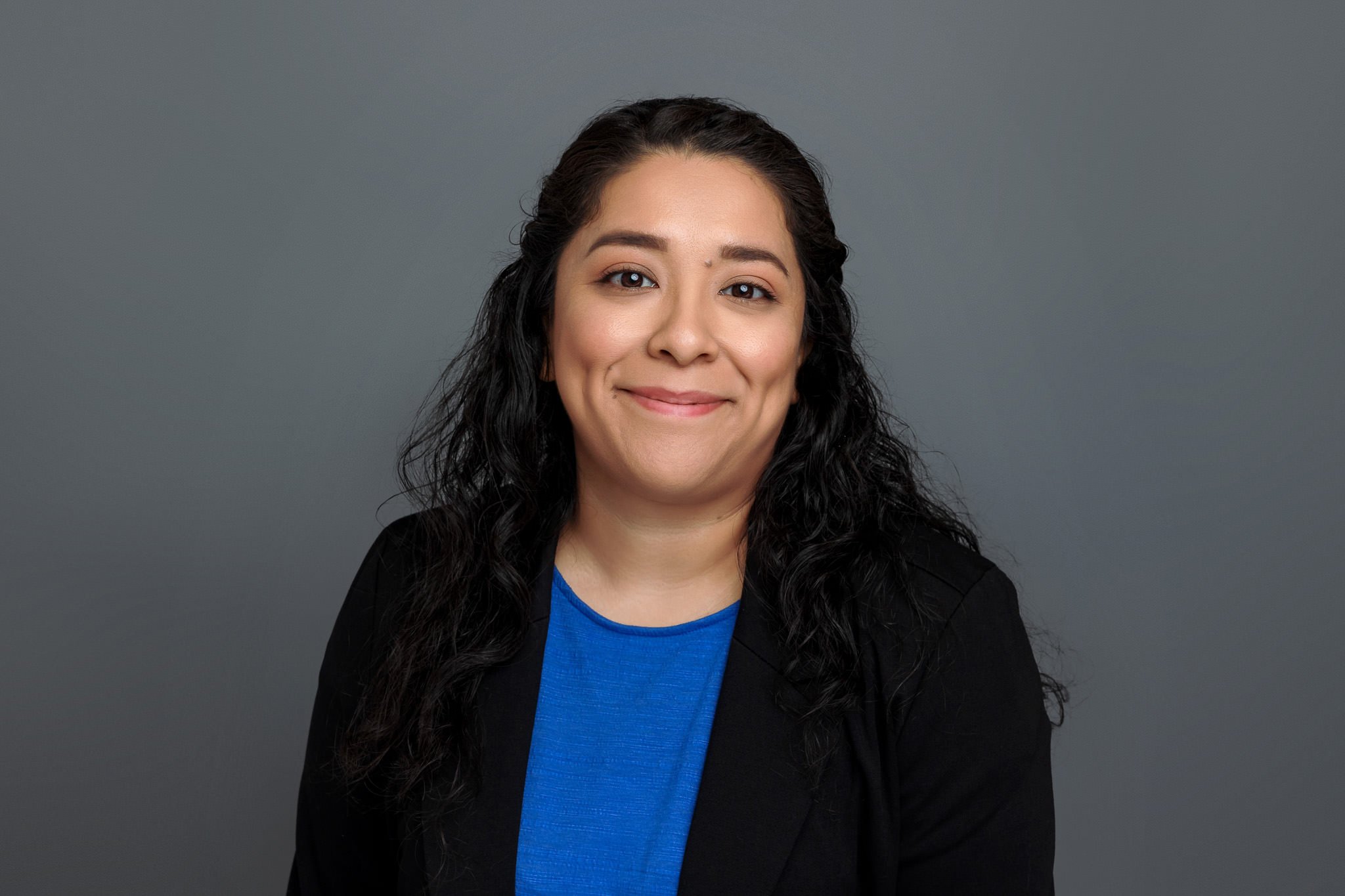 Smiling woman with long dark hair in a black blazer and blue top against a gray background.