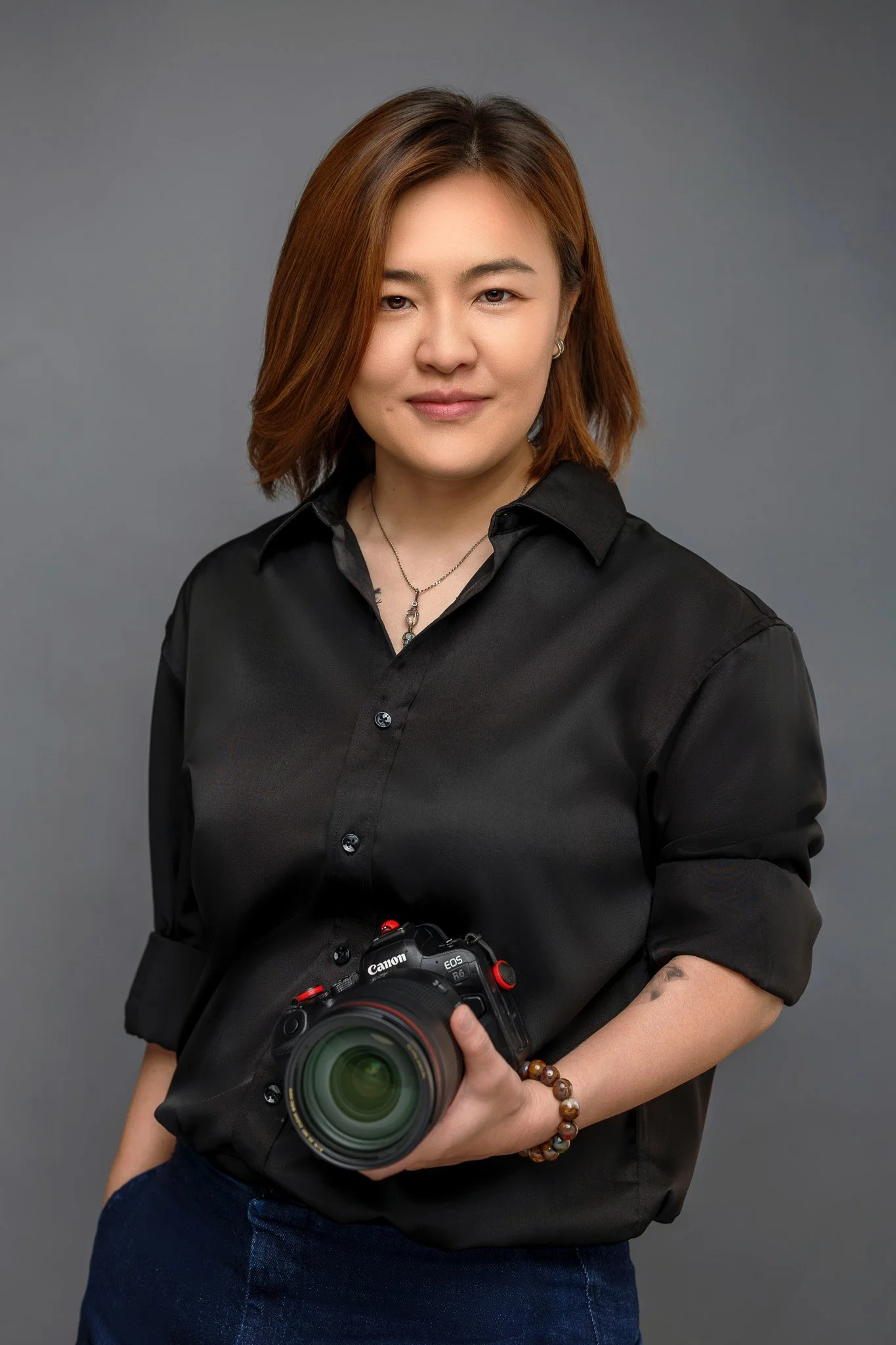 A woman with shoulder-length brown hair wearing a black shirt and holding a Canon camera with a large lens, standing against a gray background.
