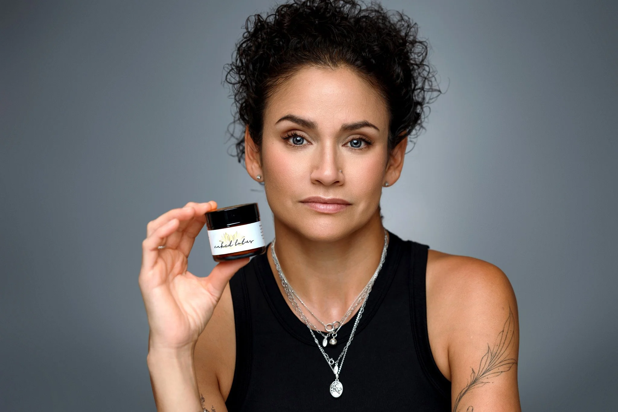 A woman with short dark curly hair and tattoos on her arm, holding a small jar of skincare product labeled 'naked balm' against a gray background.
