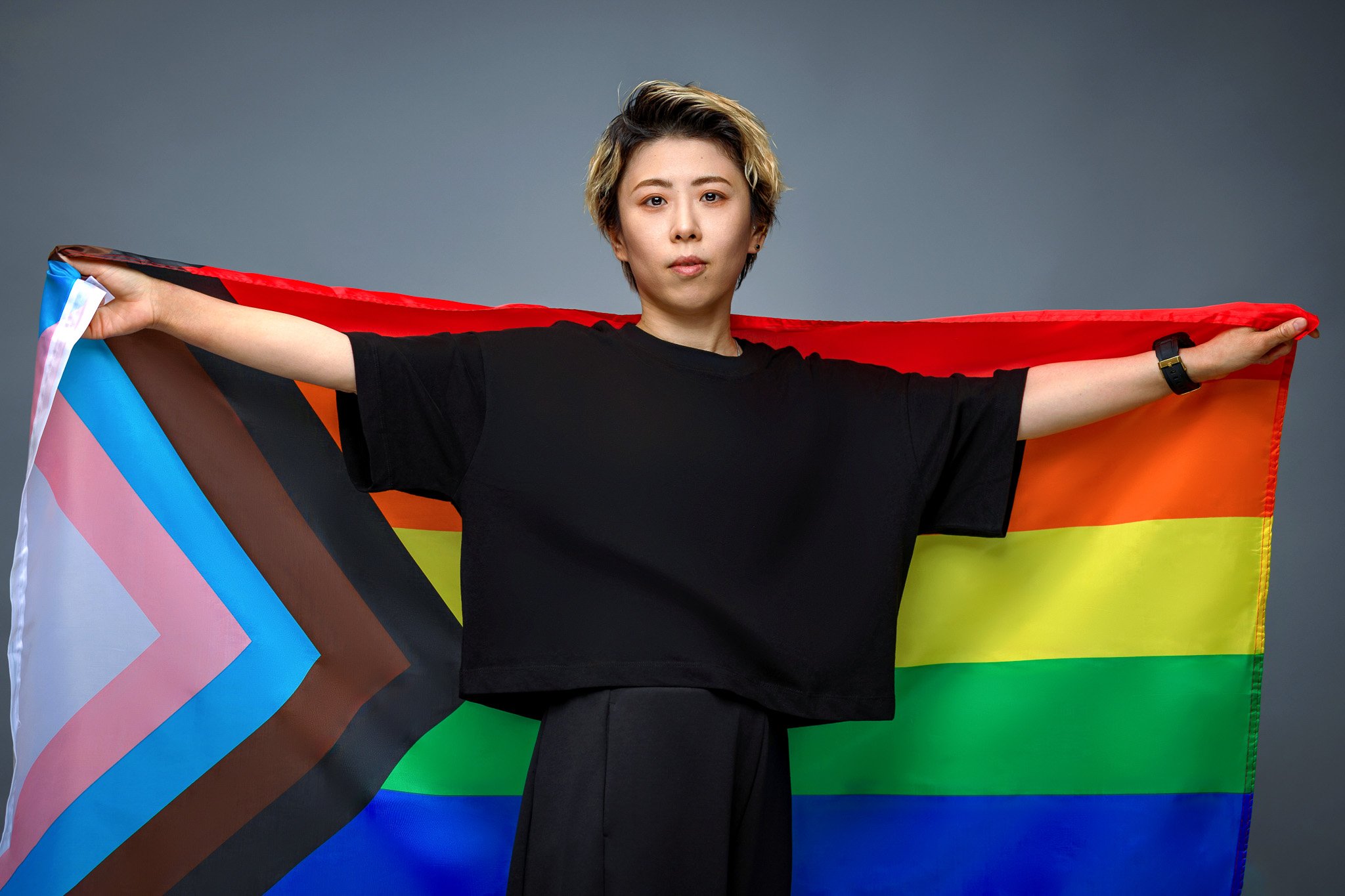 Person holding a rainbow pride flag with Black, Brown, Light Blue, Pink, White, Red, Orange, Yellow, Green, and Blue stripes, standing against a neutral gray background.