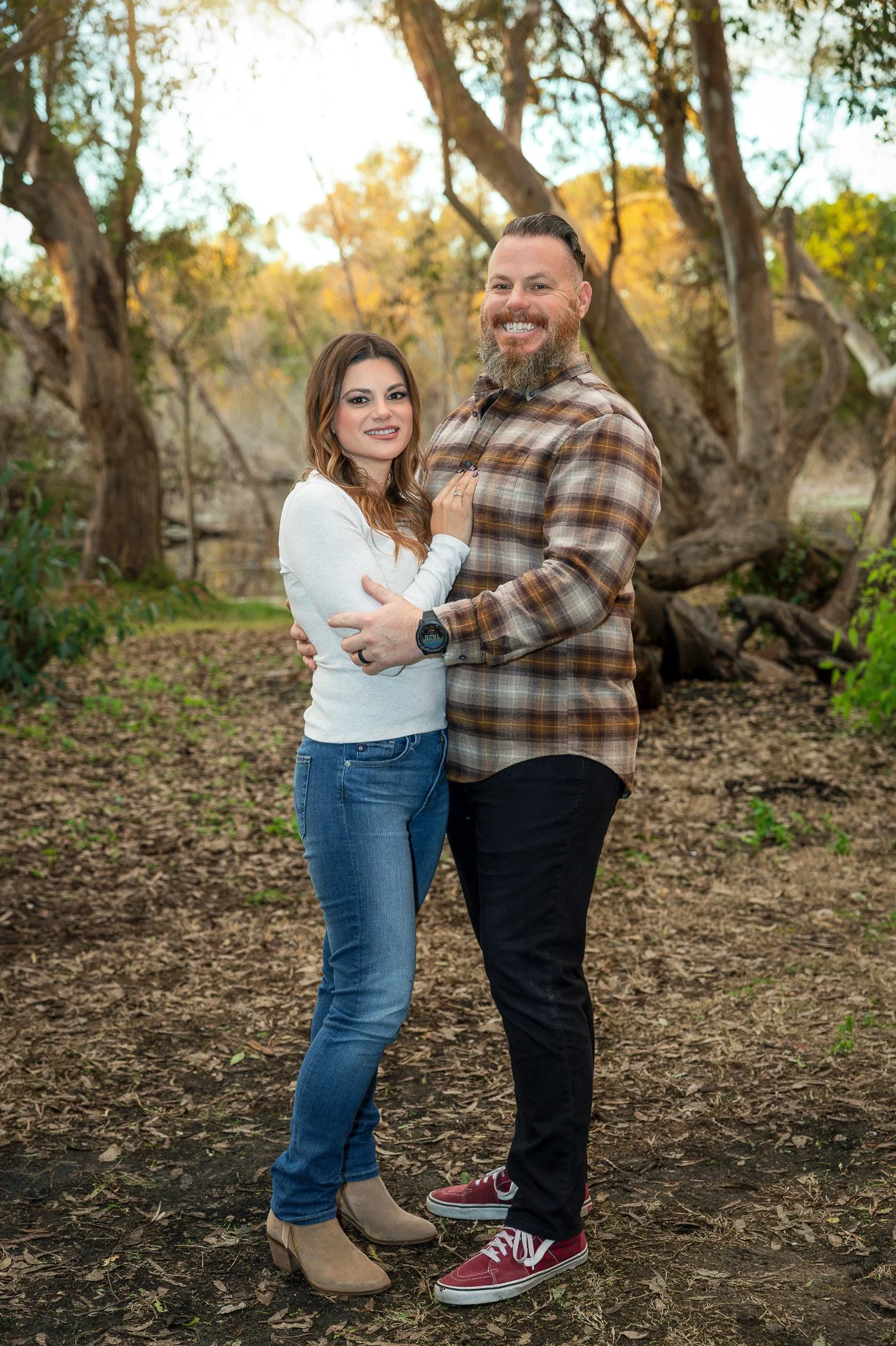A smiling couple standing close together outdoors in a forested area with trees and fallen leaves, holding hands and looking at the camera.