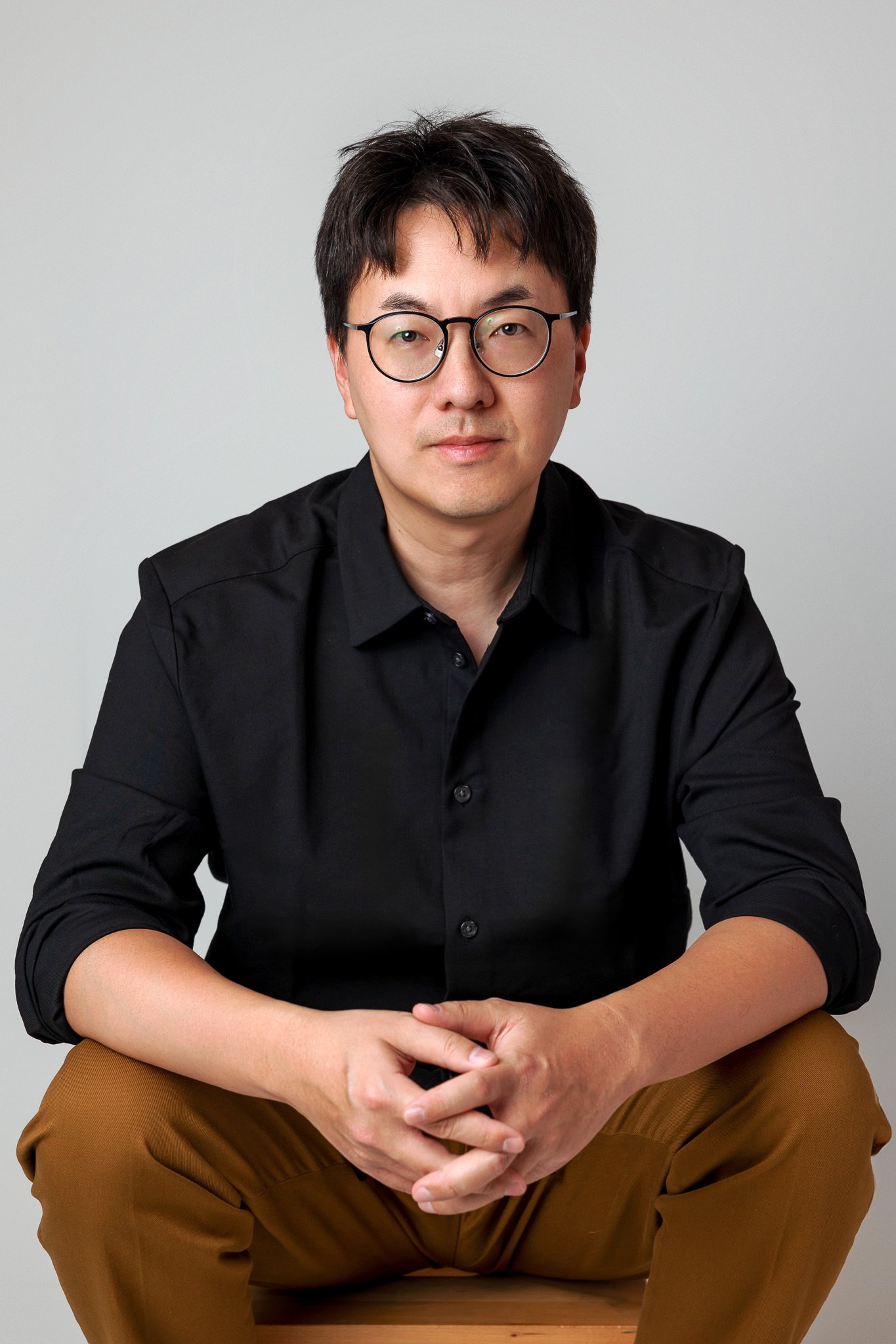 Man wearing glasses and a black shirt, sitting on a wooden stool against a white background.