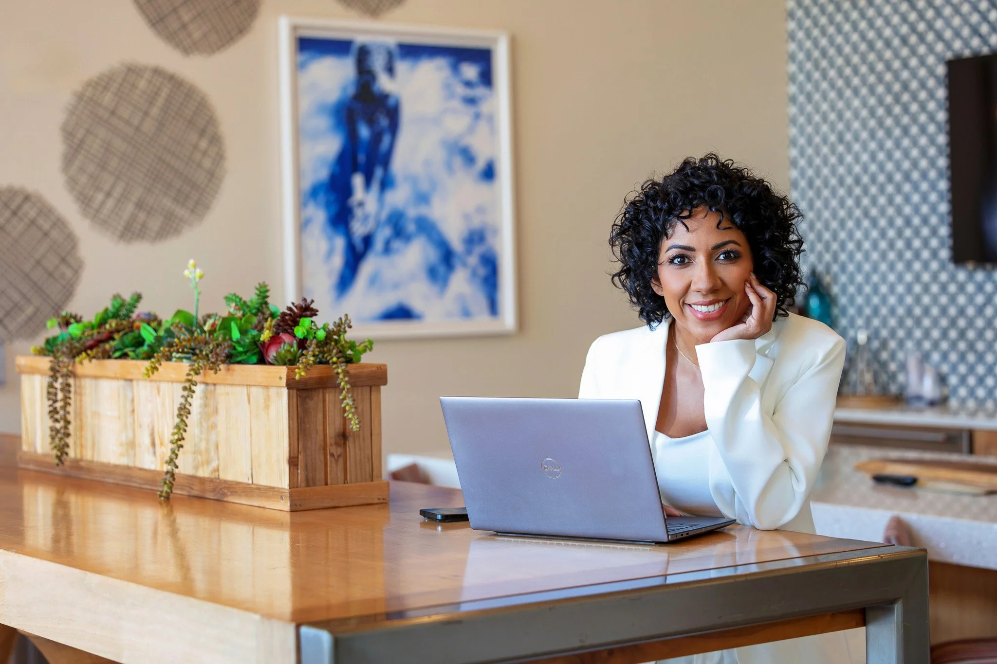 A woman with curly dark hair smiling and resting her face on her hand while sitting at a wooden table with a laptop and a smartphone. There is a wooden planter with succulents and plants on the table. In the background, there's a framed blue and whit