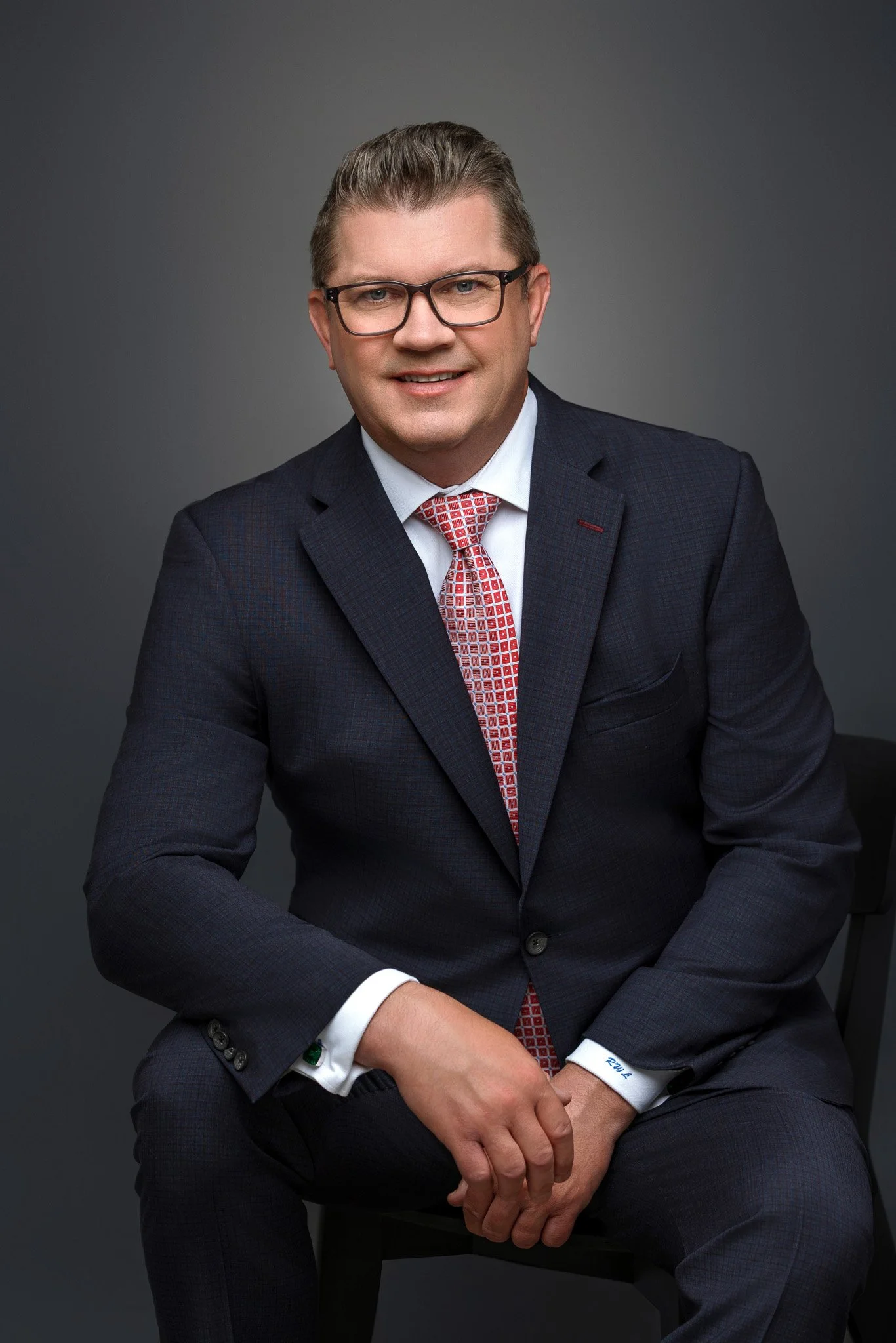 A man in a dark suit with a white shirt and red patterned tie, wearing glasses, sitting on a black chair against a gray background.