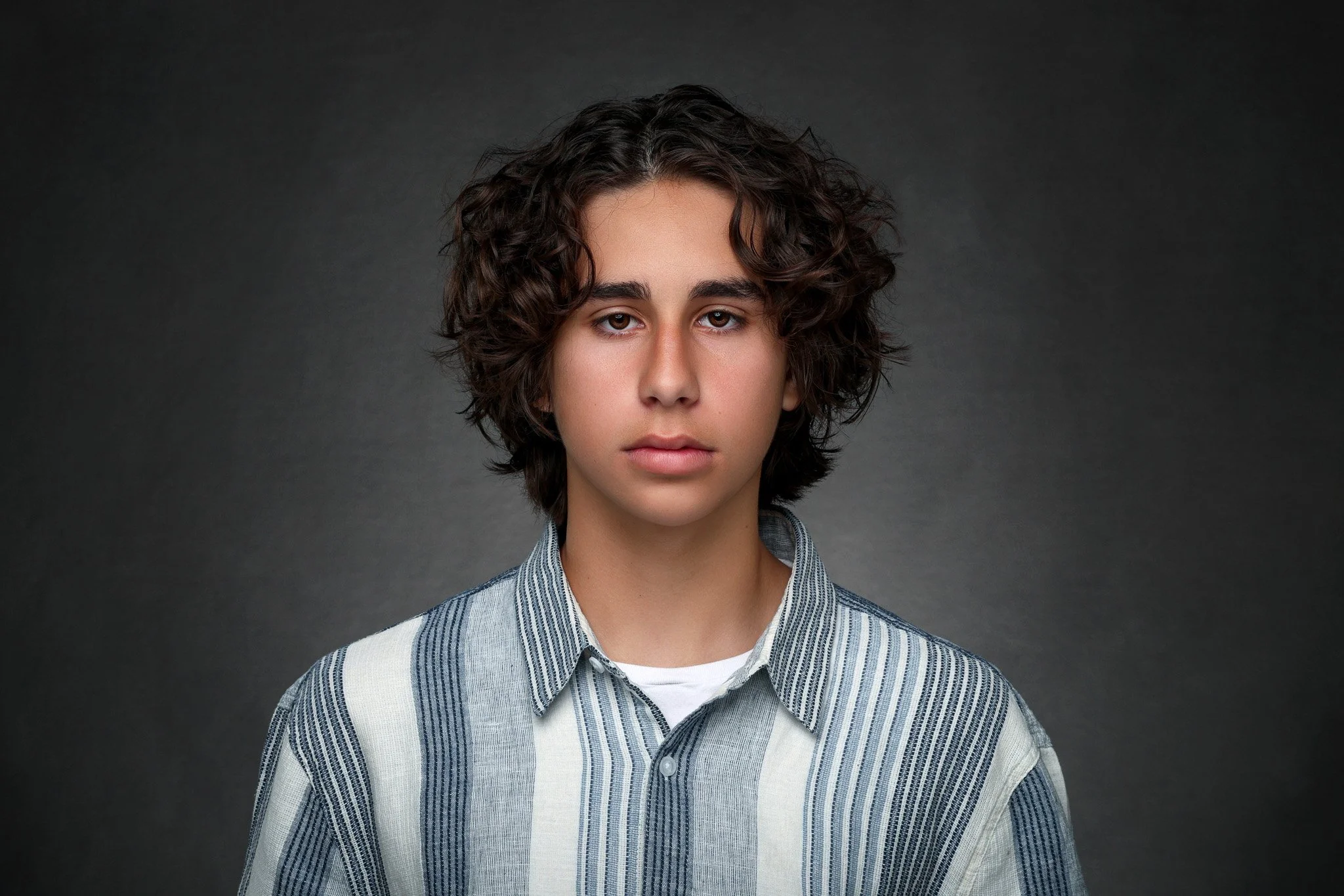 Portrait of a young person with curly brown hair wearing a striped button-up shirt and a white undershirt, against a dark gray background.