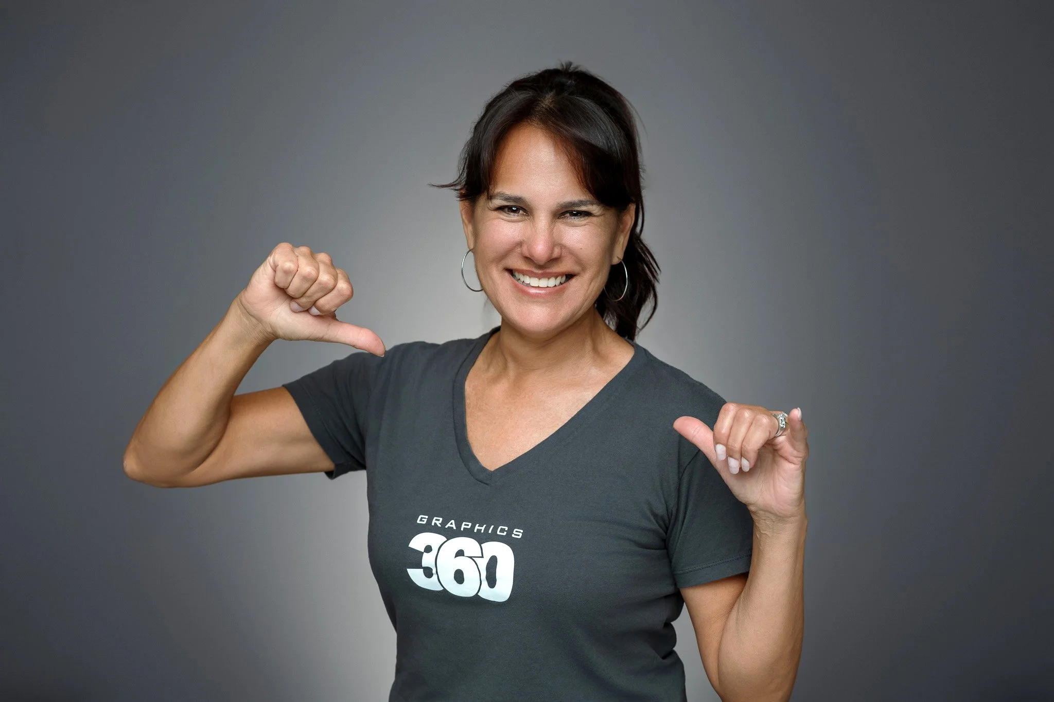 A woman in a dark gray t-shirt with 'Graphics 360' logo, smiling and pointing thumbs at her chest against a gray background.