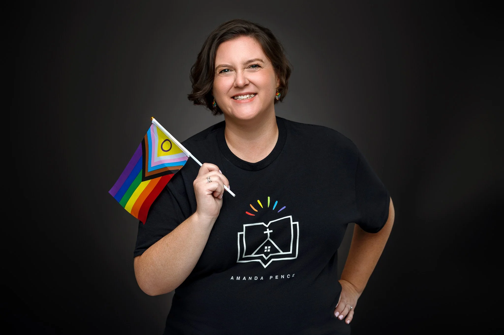 A woman with short brown hair smiling and holding a rainbow pride flag. She is wearing a black T-shirt with a colorful design and the name 'Amanda Pence' on it, against a dark background.