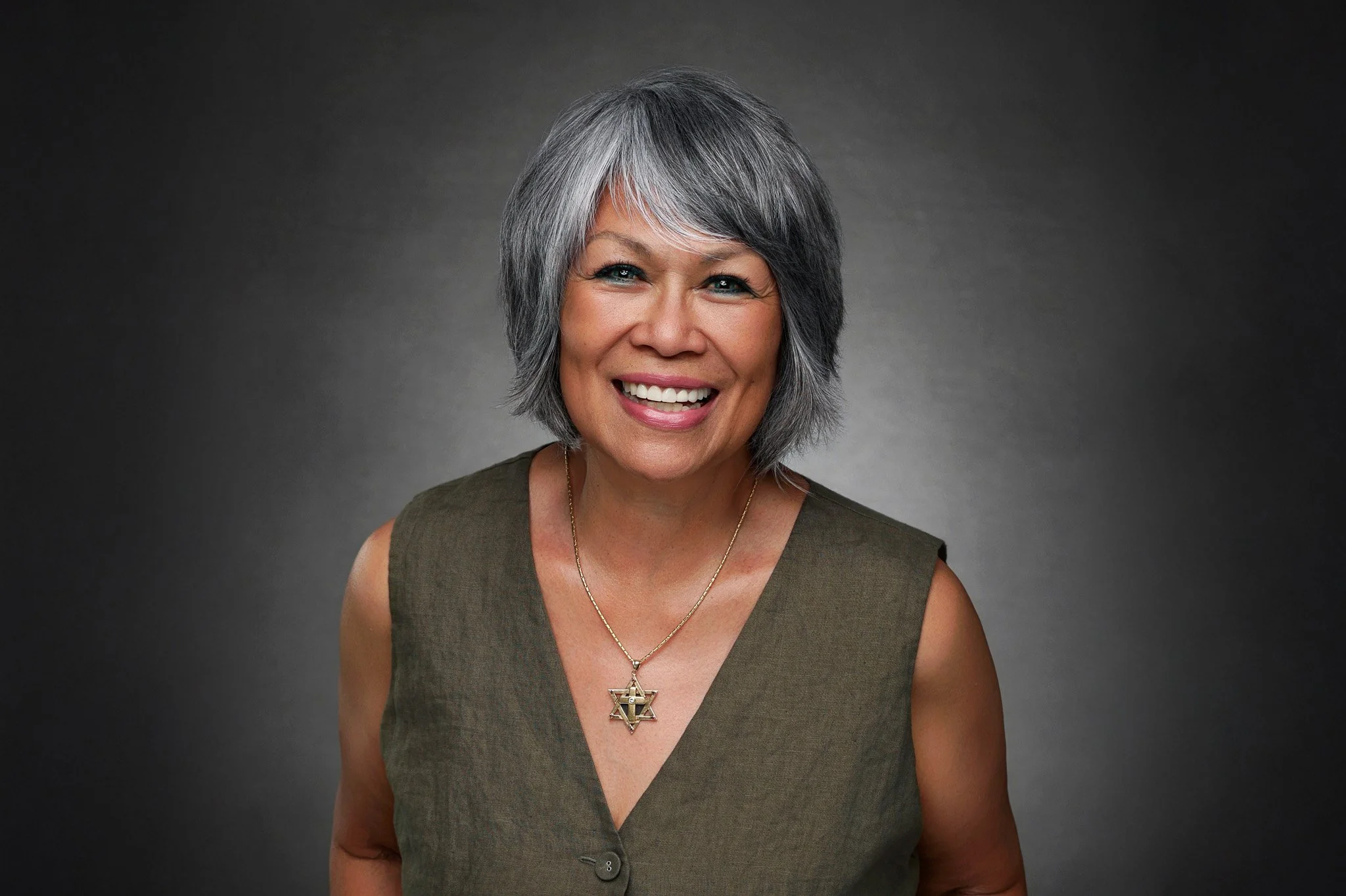 A woman with short gray hair smiling at the camera, wearing a sleeveless olive green top and jewelry with a Star of David pendant.