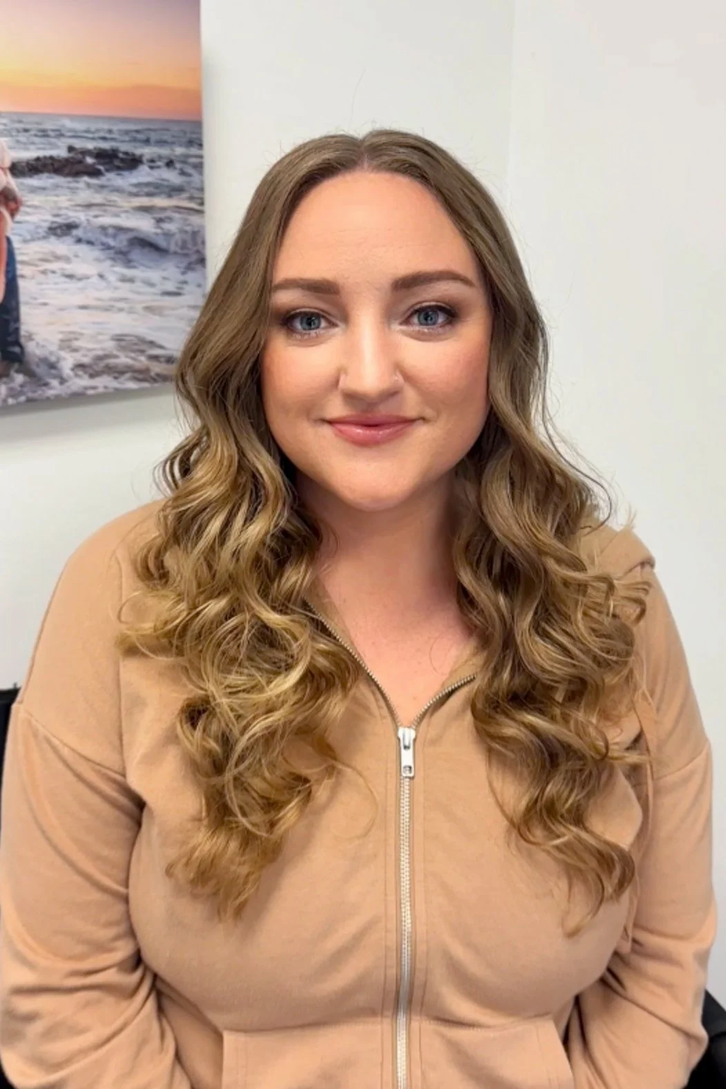 A woman with long, wavy brown hair and blue eyes looking at the camera, wearing a tan zip-up top, seated indoors with a beach sunset photo on the wall behind her.