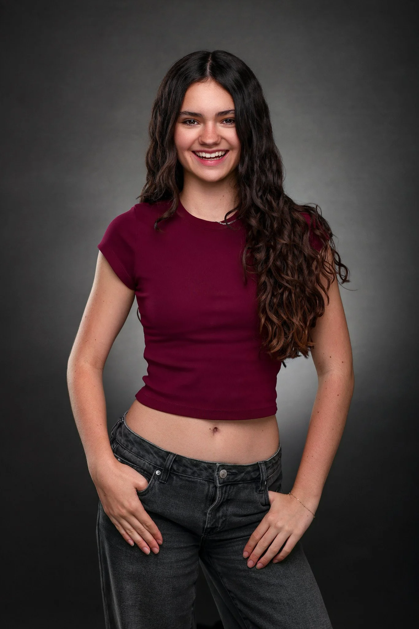 A young woman with long, curly dark hair smiling, wearing a maroon crop top and dark jeans in front of a dark grey background.