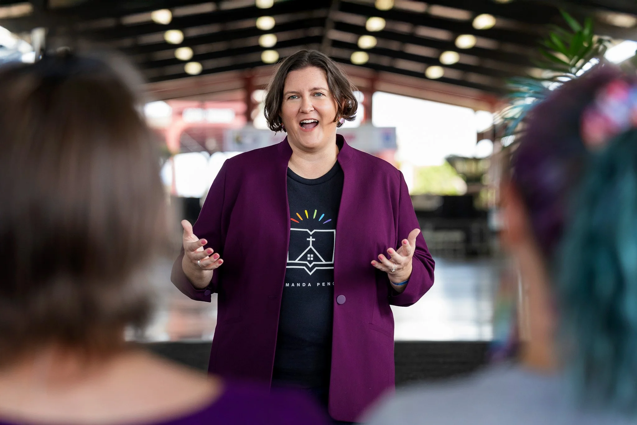 A woman with short brown hair, wearing a purple jacket and black t-shirt, speaking to a group of people in an outdoor covered area.