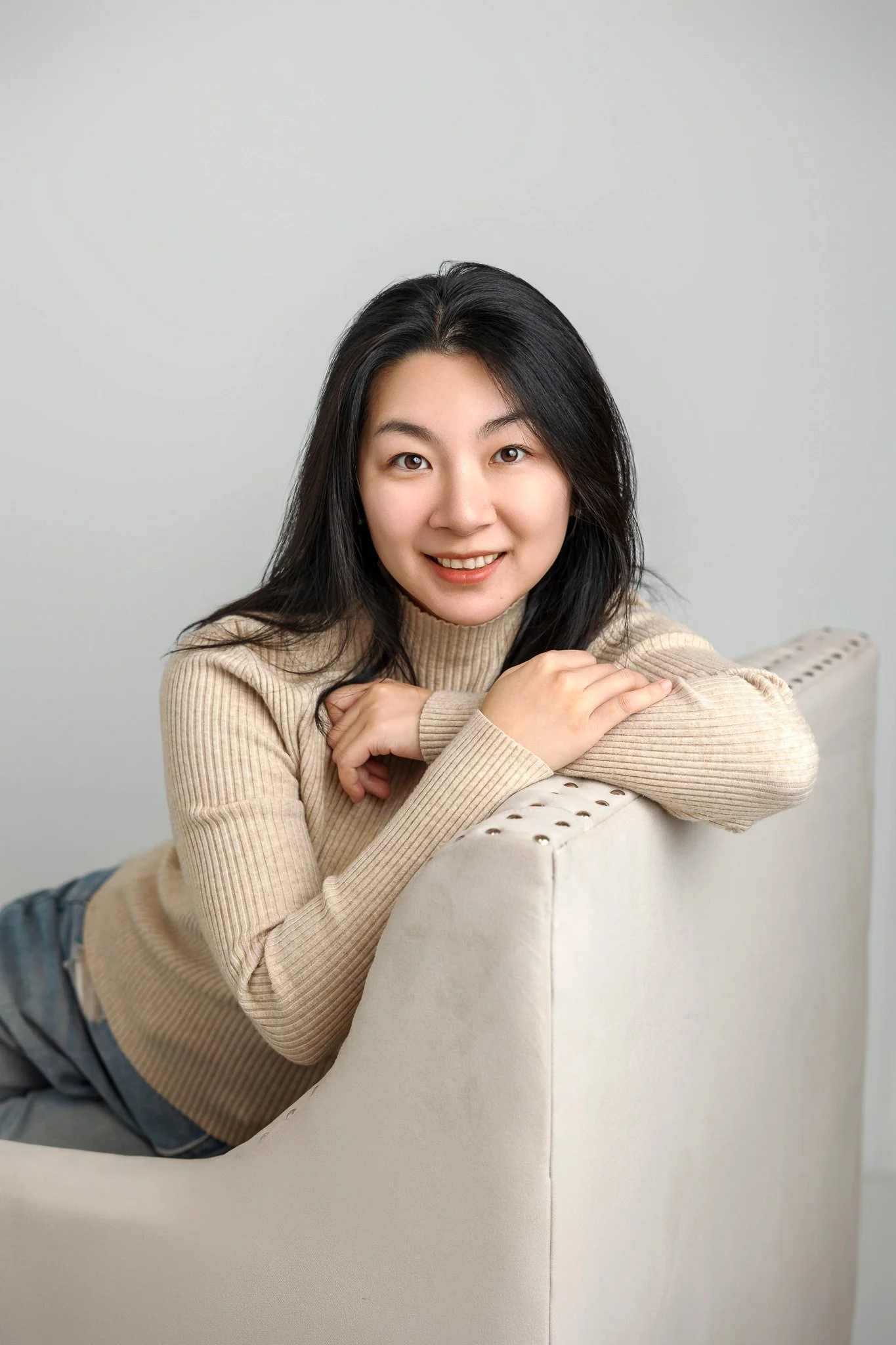 Person with long dark hair and a beige sweater smiling, leaning on a light-colored sofa.