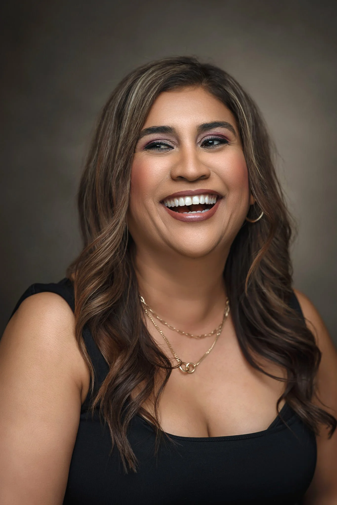 A woman with long, dark wavy hair, wearing a black top, multiple gold necklaces, and hoop earrings, smiling with her teeth visible.